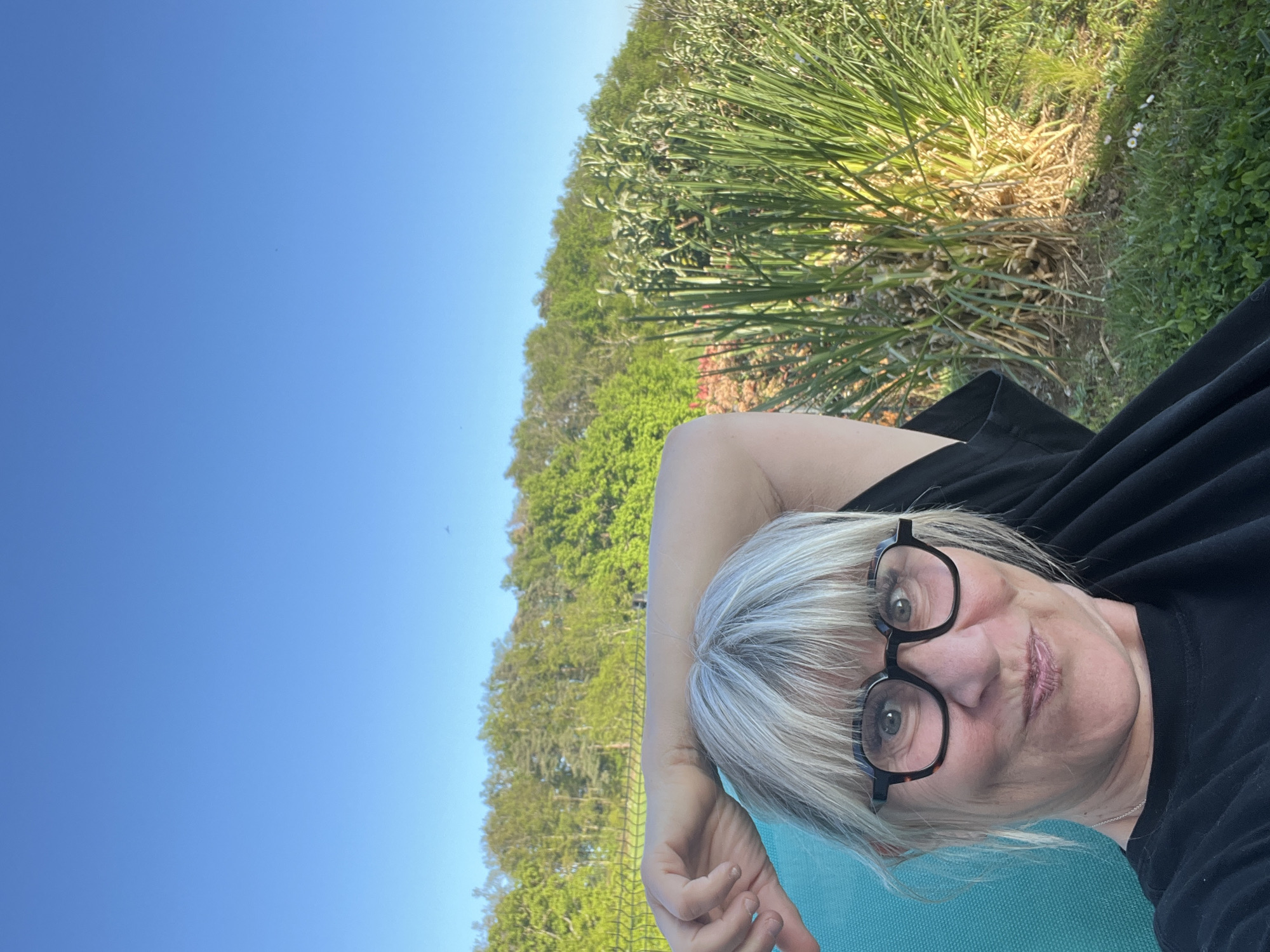 A woman with short, gray hair and glasses is lying down on a blue surface outdoors. She is wearing a black shirt. The background includes green grass, trees, and a clear blue sky. The location is Chambœuf, France.