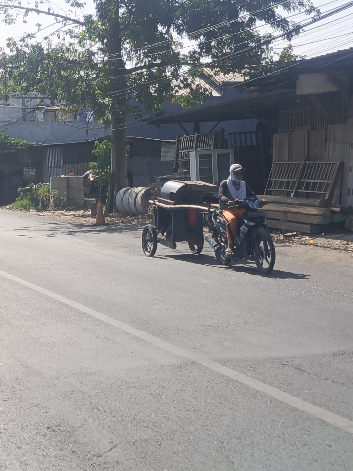 A person is riding a motorcycle with a trailer attached, moving along a paved road in Sidowungu, Indonesia. The person, wearing a light-colored head covering and orange pants, is seated on the motorcycle. The trailer is connected to the back of the motorcycle and appears to be carrying some sort of equipment or goods. The scene is outdoors, with buildings and trees visible in the background. The road is asphalt with a white line marking the lane. The lighting suggests it is daytime, with shadows cast on the road. The environment appears to be a commercial or mixed-use area, with what looks like a workshop or store with materials and windows on the right side of the image.
