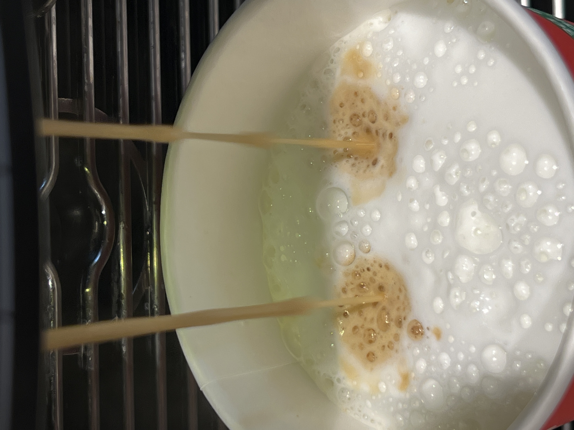 The image shows a top-down view into a white paper cup filled with a milky beverage, likely coffee or a similar drink, containing foam and patches of espresso. Two streams of a dark liquid, presumably espresso, are being poured into the cup from the left side of the frame. To the left of the cup, parts of a metal coffee machine are visible. The image provides no information about the city of Krasnodar or Russia.