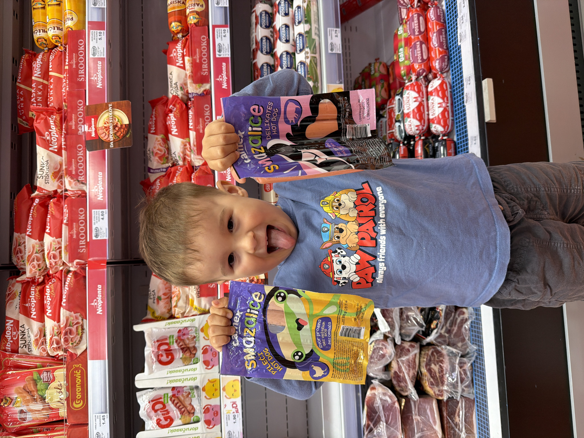 A young, joyful child is pictured in a supermarket aisle in Budva, Montenegro, holding up two packages of hot dogs with a playful expression, his tongue extended. The child, likely a boy with light brown hair, wears a blue t-shirt adorned with Paw Patrol characters (Chase, Marshall, Rubble, Skye) and the phrase "Always friends with everyone."
He holds two "Smazalice Delikates Hot Dog" packages; one is purple and pink, the other yellow and green. Visible text on the packaging includes "DELIKATES HOT DOG," "PILEĆI HOT DOG" (chicken hot dog), "izvor proteina" (source of protein), "bez veštačkih boja" (no artificial colors), "bez omotača" (no casing), "205 g," and an expiration date of "07/07/2026."
The background reveals a well-stocked refrigerated section of a grocery store. Numerous packaged meat products are displayed on shelves, predominantly featuring "Neoplanta" brand items like "ŠIROOOKO" (salami), "SUNKA STIŠNJENA" (pressed ham), "CUREĆA PRSA" (turkey breast), and "PILEĆA PRSA" (chicken breast). A promotional sign reads "DA SPOJIMO STOLOVE" (let's connect tables). Other brands visible include "Goranović" with "VIRŠLA VAKUM" (vacuum-packed wieners) priced at €1.45, and "GUDI" brand sausages with "doručaaak!" (breakfast!) priced at €1.90. Shelves also contain "POSEBNA KOBASICA" (special sausage) and "Gala" products, with prices such as €5.10, €4.60, and €2.75 visible. The labeling predominantly in Serbian/Croatian aligns with the Montenegrin context. The image captures a lively moment during a shopping trip.