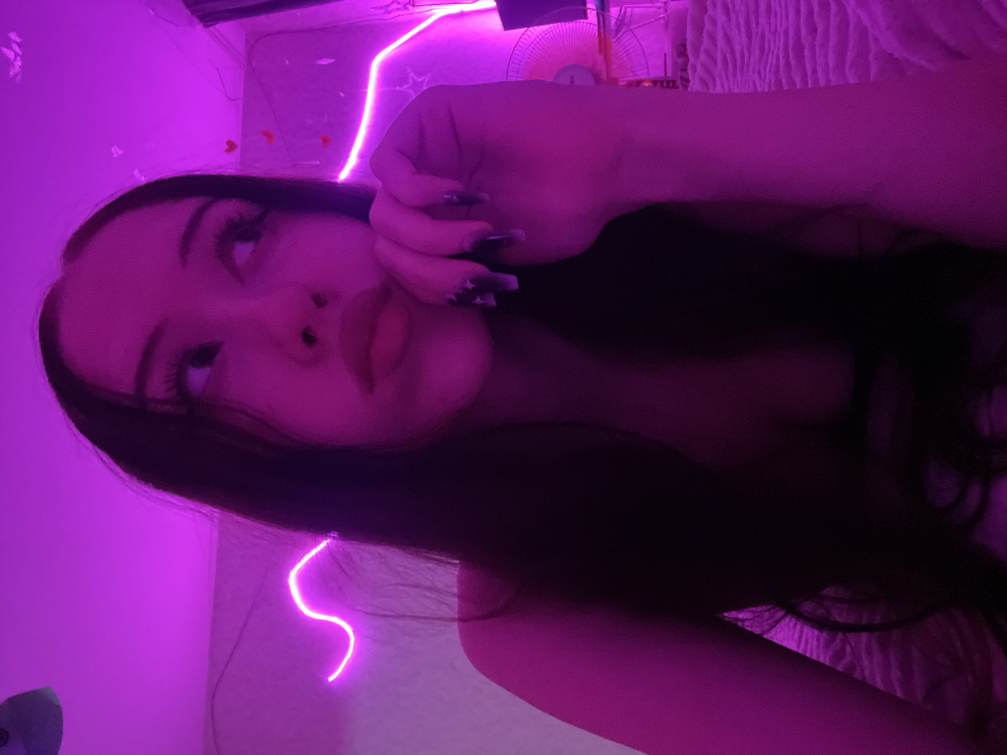 The image shows a young woman with long dark hair illuminated by purple lighting. She is resting her chin on her hand, looking away from the camera. Her nails are painted dark. In the background, there are pink neon lights and some small heart-shaped decorations on the wall. A fan is visible to the right. The overall environment suggests a bedroom or a similar personal space. No specific details about Votkinsk, Russia, are discernible from the image itself.