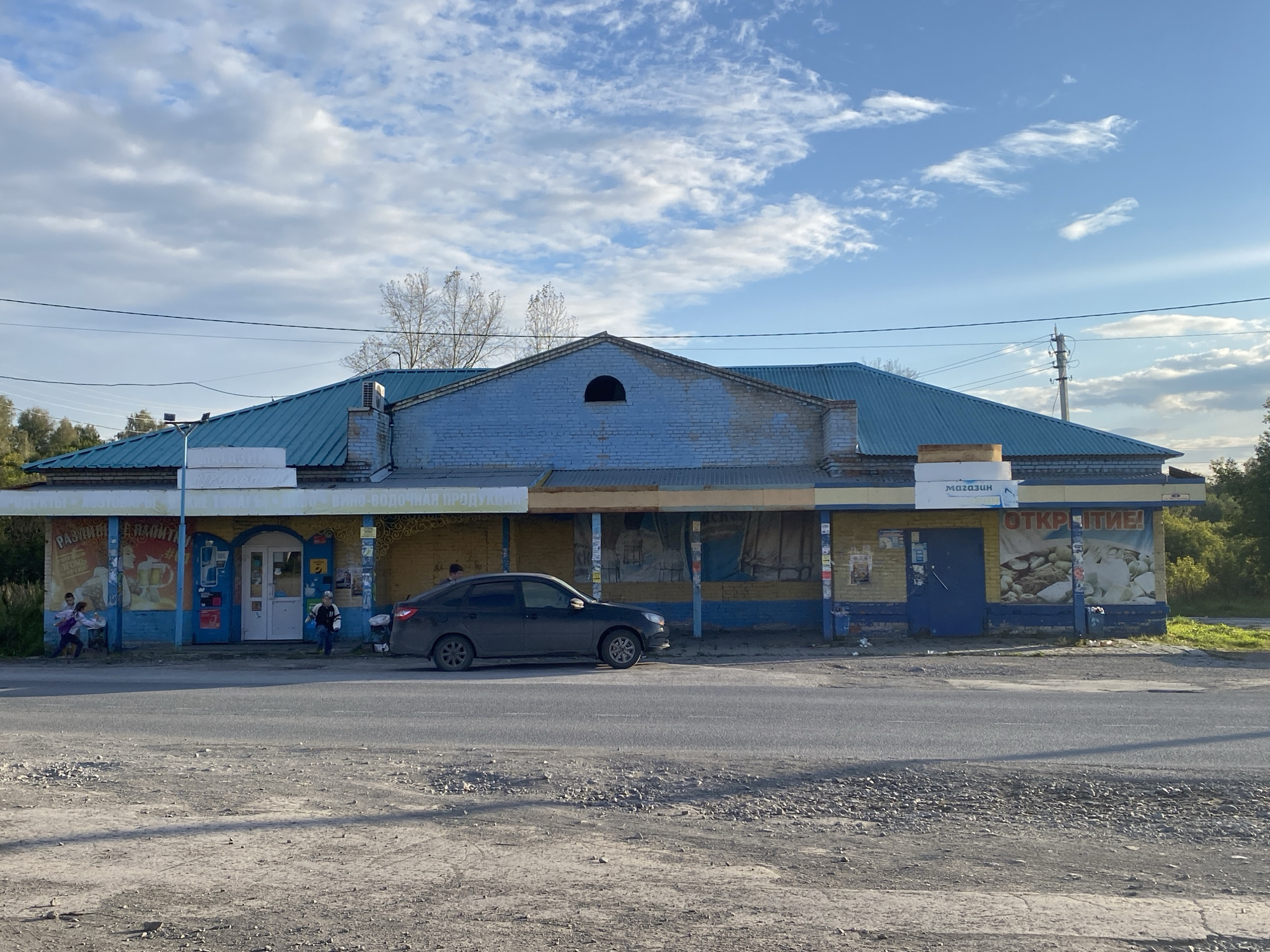 The image depicts a single-story, blue and yellow building with a corrugated metal roof, situated in Listvyanskiy, Russia. The building appears to be a shop or small store, with several signs visible, including one that reads "МАГАЗИН" (Magazin - Store) and another that says "ОТКРЫТИЕ!" (Otkrytiye! - Opening!). There are posters on the exterior walls. A dark-colored sedan is parked in front of the building. Several people are visible: two children and an adult are near the left side of the building, and a child is near the center of the building. The building is located on a paved road with a gravel shoulder in front. Trees and foliage are visible in the background, and the sky is partly cloudy.