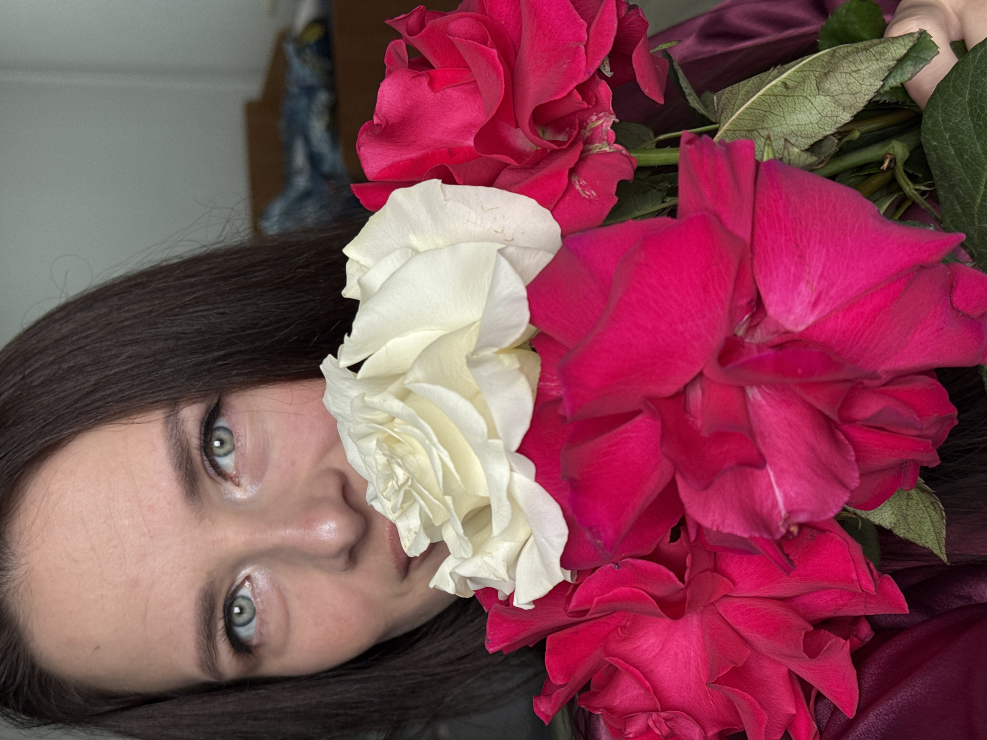 A close-up, eye-level medium shot captures a person holding a bouquet of roses. The person, with dark hair and light eyes, is partially obscured by the flowers, only their eyes and mouth are visible. They are wearing a dark red garment that appears to be satin. The bouquet consists of several large roses, with two prominent red roses in the foreground and a white rose positioned above them. The background is blurred and indistinct, showing a plain wall and what appears to be furniture. The scene is indoors. The image does not provide any specific information that would allow for localization in the city of Rusalevka, Russia.