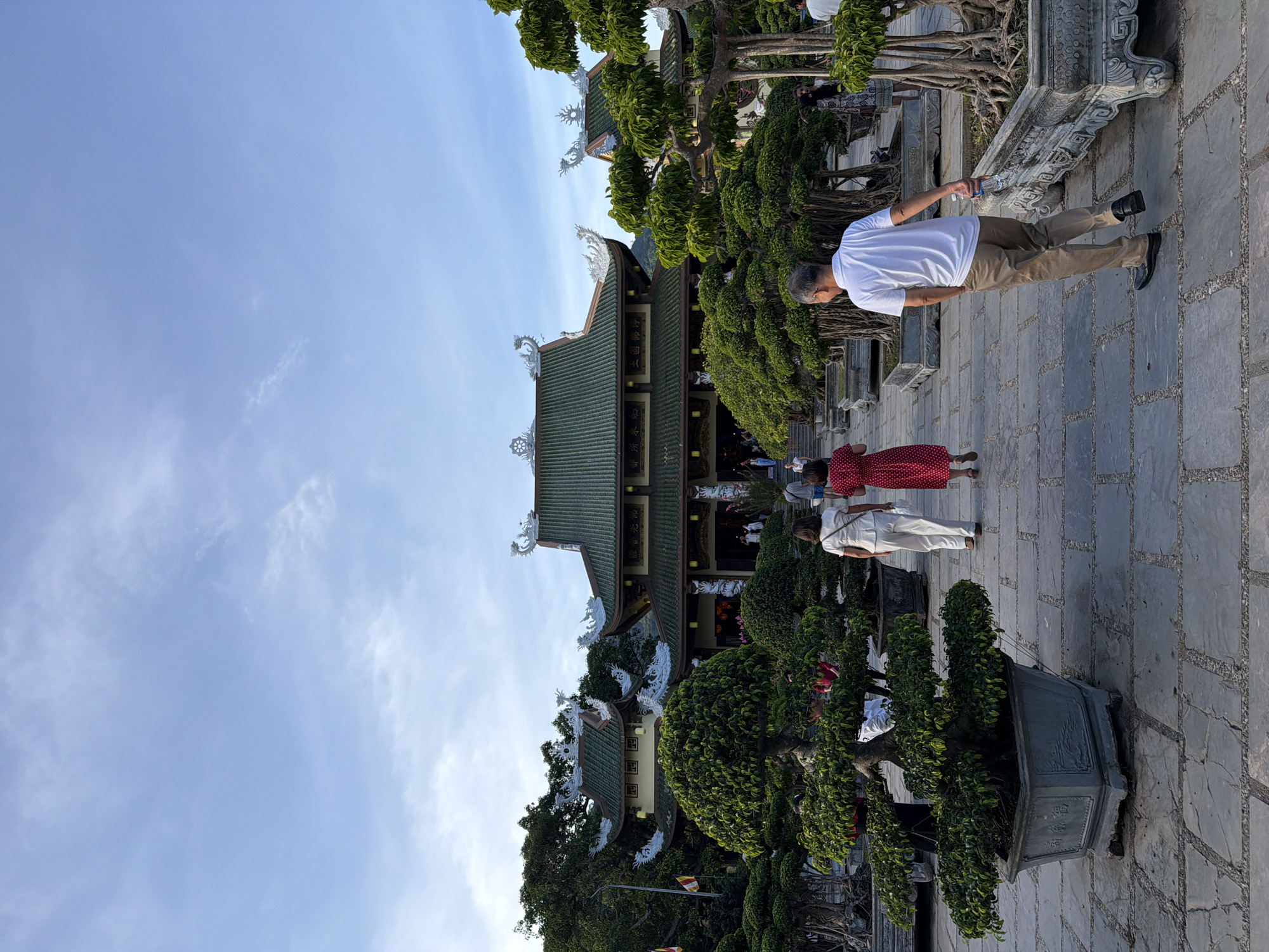 The image shows a view of a temple in Da Nang, Vietnam, featuring traditional architecture with green tiled roofs and decorative dragon motifs. In the foreground, a paved walkway is lined with large, meticulously shaped bonsai trees in stone planters. Several people are walking on the path, heading towards the temple entrance. One man in a white polo shirt and tan pants walks away from the camera, holding a bottle of water. Two women in white and red dresses are walking ahead of him, also heading towards the temple. The sky above is a pale blue with some scattered clouds, and lush green trees surround the temple complex. The overall scene suggests a serene and well-maintained religious or historical site.