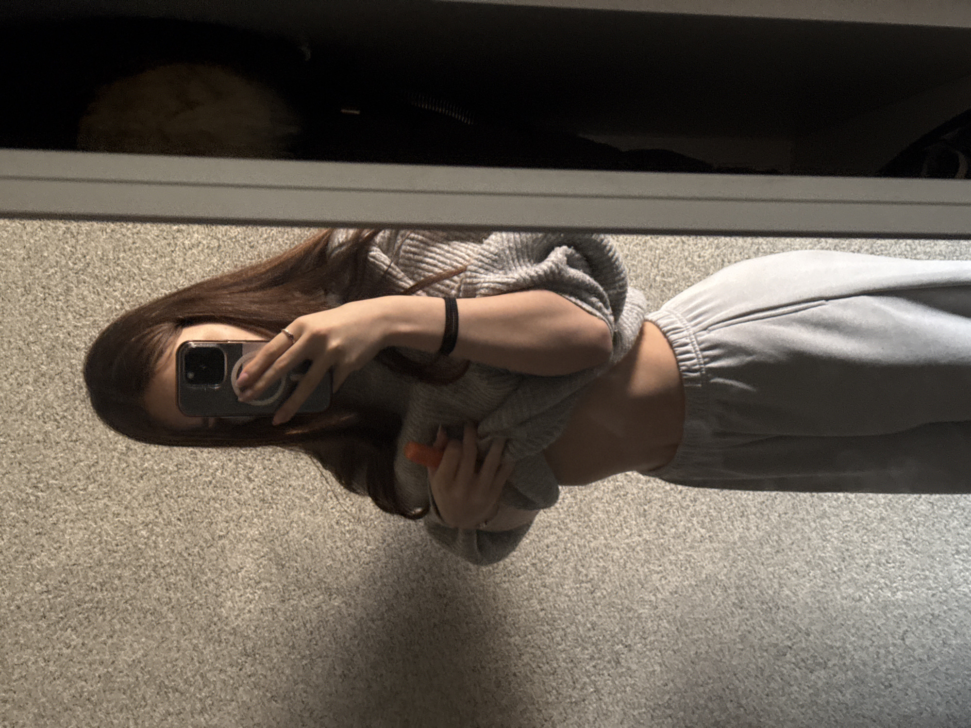 A woman is taking a mirror selfie in what appears to be a closet or storage area. She is wearing grey sweatpants and a grey sweater that is slightly pulled up, revealing part of her torso. She holds a dark-colored smartphone in her right hand to capture the image. Her hair is long and brown. The background includes a carpeted floor, a shelf or ledge above the mirror with items stored on it, and part of her reflection in the mirror. There is no information to suggest this picture was taken in Sofia, Bulgaria, or any other specific location.