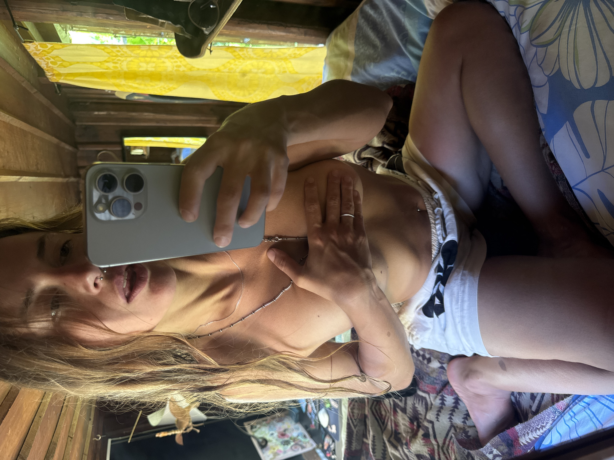 The image shows a young woman with long, light brown hair, a nose ring, and fair skin, taking a selfie in what appears to be a rustic, wooden-paneled room. She is sitting cross-legged on a bed with patterned blankets. She is wearing white shorts with a black bow emblem. Her arms are crossed in front of her chest, partially covering her bare torso. A silver necklace with small beads and a pendant hangs around her neck. In the background, a bright yellow curtain with a patterned design is visible behind a window, and to the left, a mirror reflects a part of the room. The lighting suggests it might be daytime, with natural light coming from the window. The overall setting appears casual and personal.