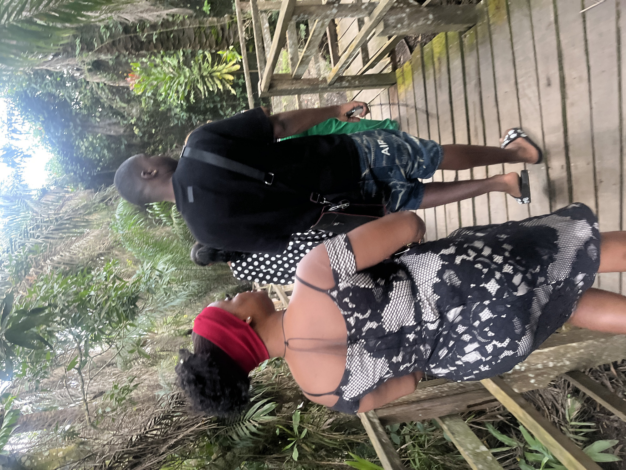 In Lekki, Nigeria, a group of people walk along a wooden boardwalk amidst dense foliage. A woman in the foreground, wearing a black and white lace dress and a red headband, walks away from the camera. To her right, a man in a black t-shirt and denim shorts walks ahead. Behind them, another individual in a black and white polka dot dress and a person in a green outfit are visible. The boardwalk is elevated, suggesting a path through a swampy or overgrown area, possibly a nature reserve or park. The lighting suggests an overcast day.