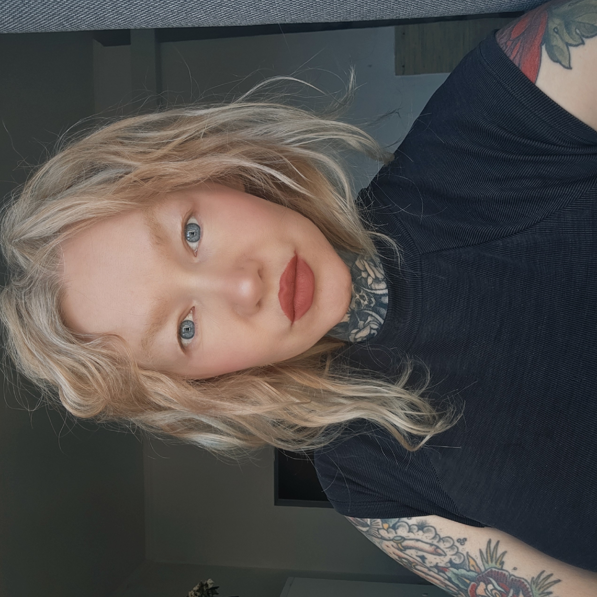 A woman with light blonde, shoulder-length wavy hair, fair skin, and blue eyes is facing the camera. She has a neutral expression with rosy cheeks and red lipstick. She is wearing a black, ribbed top. Tattoos are visible on her neck and arm. The background includes a gray textured surface, likely a couch, and a dark gray wall with a hint of white trim near the bottom. The presence of a television screen mounted on the wall suggests an indoor setting, possibly a living room. The location is not identifiable as Saint Petersburg, Russia, based on the available visual information.
