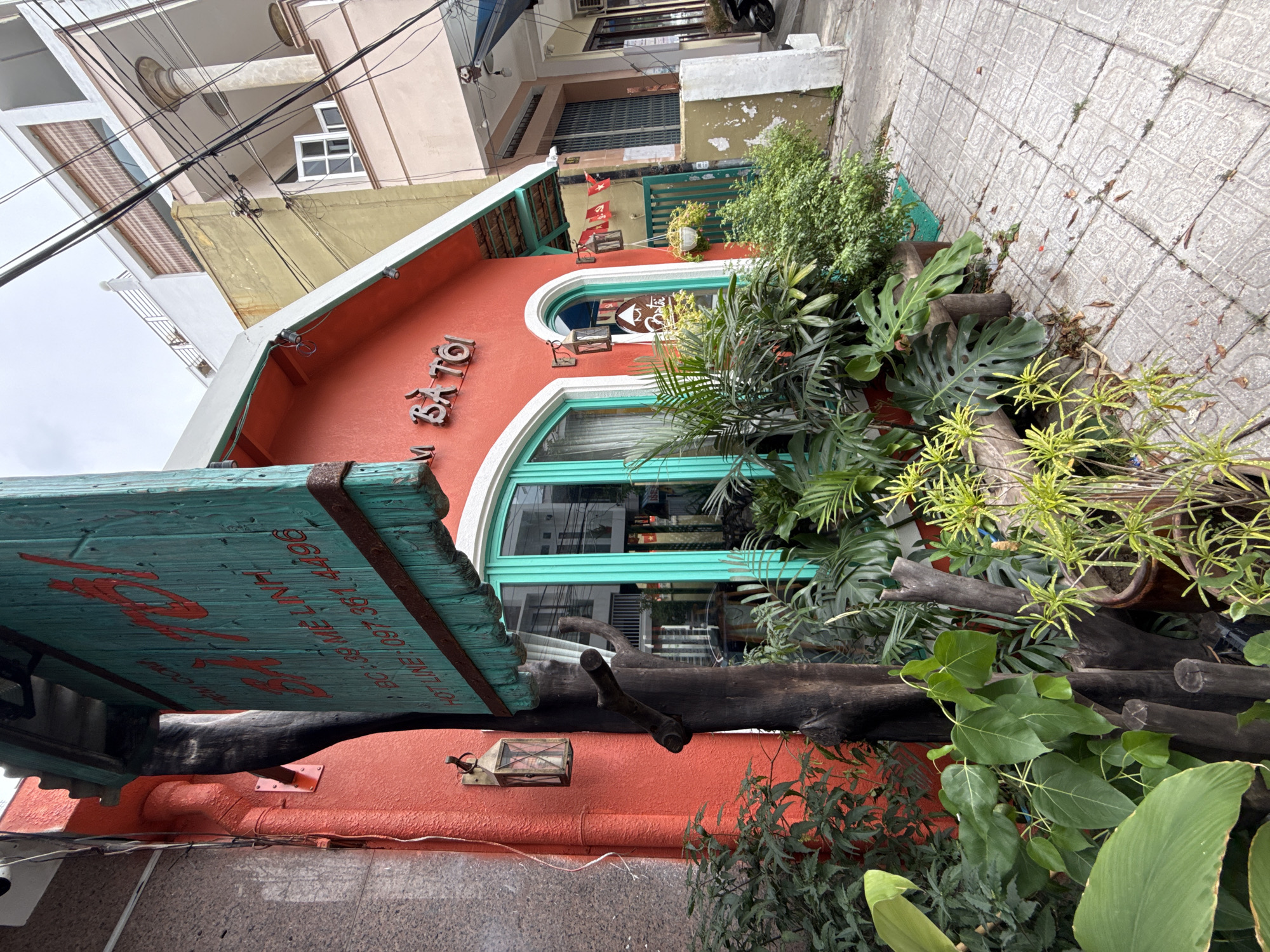 The image depicts the exterior of a red building with turquoise-trimmed arched windows and doors in Nha Trang, Vietnam. A wooden sign with red lettering hangs from the building. Text on the sign includes "BA TOI," "BC.39 ME LINH," and "HOT LINE: 097.361.4496." Lush green plants, including large-leafed tropical plants and smaller shrubs, are situated in front of the building, some in pots. Several dark brown, rough-textured wooden posts are also visible in the foreground, integrated with the plant life. The building is situated on a tiled sidewalk. In the background, other buildings are visible, along with overhead electrical wires. Small red flags with yellow stars hang from the side of the building near an entrance. The overall scene suggests a commercial establishment, possibly a restaurant or shop, with a focus on a natural or organic theme.