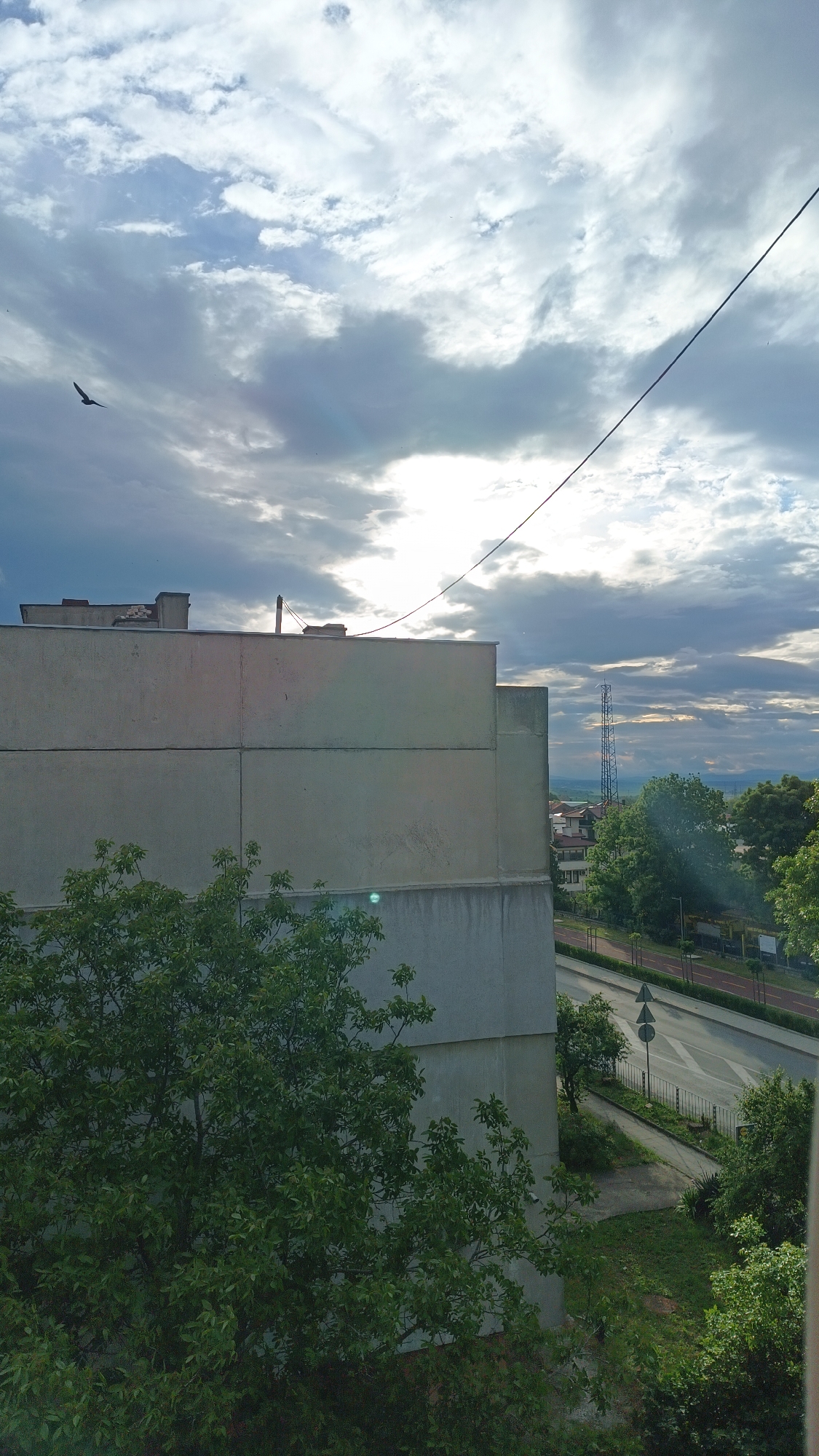 The image depicts an exterior urban scene with a focus on a large, drab concrete building in the foreground. The building is multi-storied, with a flat roof and simple, modular construction. The exterior of the building appears weathered and slightly stained. In front of the building is a dense green tree, partially obscuring the structure.
Beyond the building, a street with light traffic is visible, alongside a pedestrian walkway. Trees line the street, providing a green contrast to the built environment. In the distance, a tall communications tower stands on the horizon, behind rolling hills and a hazy sky.
The sky itself is overcast with textured clouds, and there is a bird flying in the left upper corner. The light suggests it is daytime, but the sun is obscured.
The location is likely a residential area, but without more information, the specific location within the city of Unknown, Unknown cannot be determined.