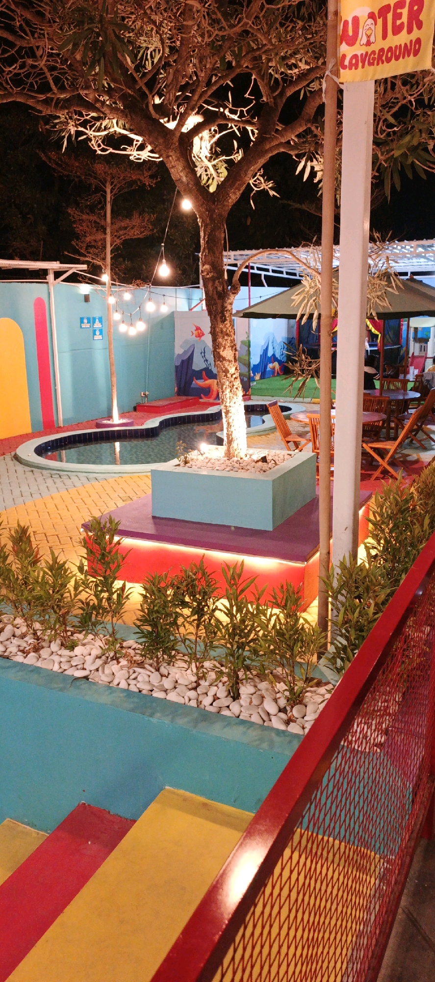 The image depicts a vibrant, outdoor recreational area at night, likely a playground or entertainment venue, with a sign overhead reading "WATER PLAYGROUND". A shallow, irregularly shaped pool with dark blue water is a central feature, surrounded by colorful, painted walls adorned with underwater-themed murals, including fish and starfish. String lights are suspended above the pool, casting a warm glow. The ground is paved with a mosaic of yellow, orange, and purple tiles. A large tree with bare branches stands in the center, its trunk illuminated from below by pink and red lights. A light blue planter box filled with white pebbles and small green plants is situated near the tree. In the background, there are outdoor seating areas with wooden tables and chairs, and umbrellas providing shade. A bright red metal fence runs along the foreground, with a section of colorful stairs painted in red and yellow leading down from the left. The scene suggests a lively and engaging atmosphere, designed for enjoyment. The location is identified as Sidoarum, Indonesia.