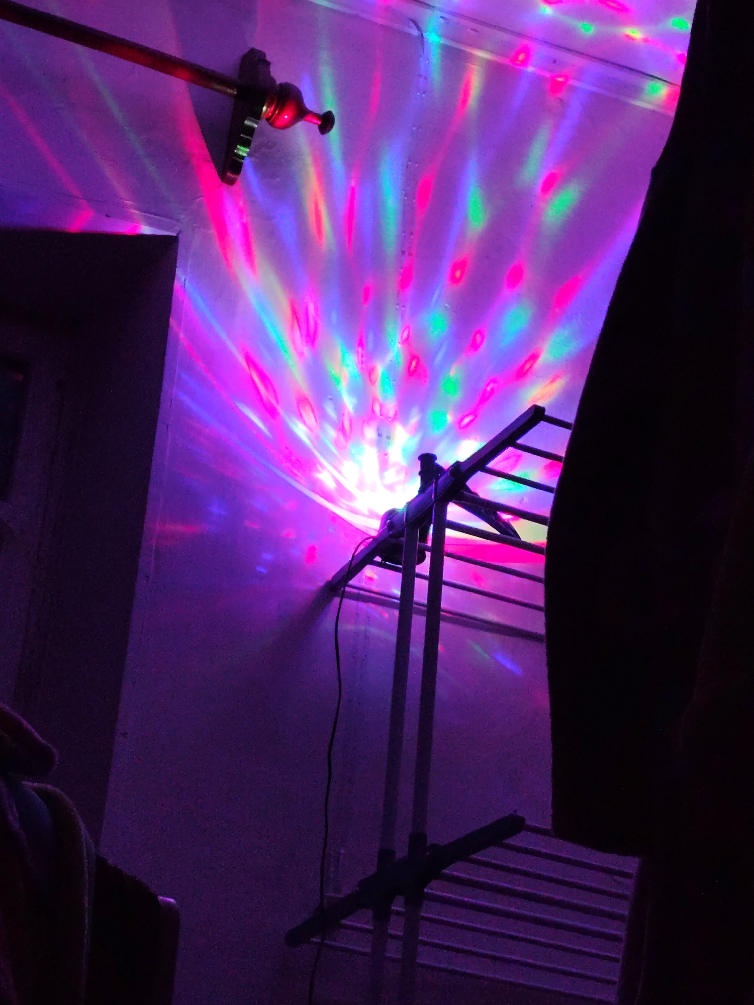 The image depicts a room illuminated by a disco ball, casting colorful lights onto the walls and a drying rack. The room appears to be located in Aksukent, Kazakhstan. A curtain rod with a decorative finial is visible on the left side of the image, with pink and purple lights reflecting off it. A metal drying rack stands in the center of the frame, with the disco ball positioned above it, projecting beams of pink, green, and blue light. A dark, silhouetted figure is partially visible on the right side of the image, obscuring a portion of the background. The walls are textured and painted in a light color, with the disco lights creating a vibrant, kaleidoscopic effect.