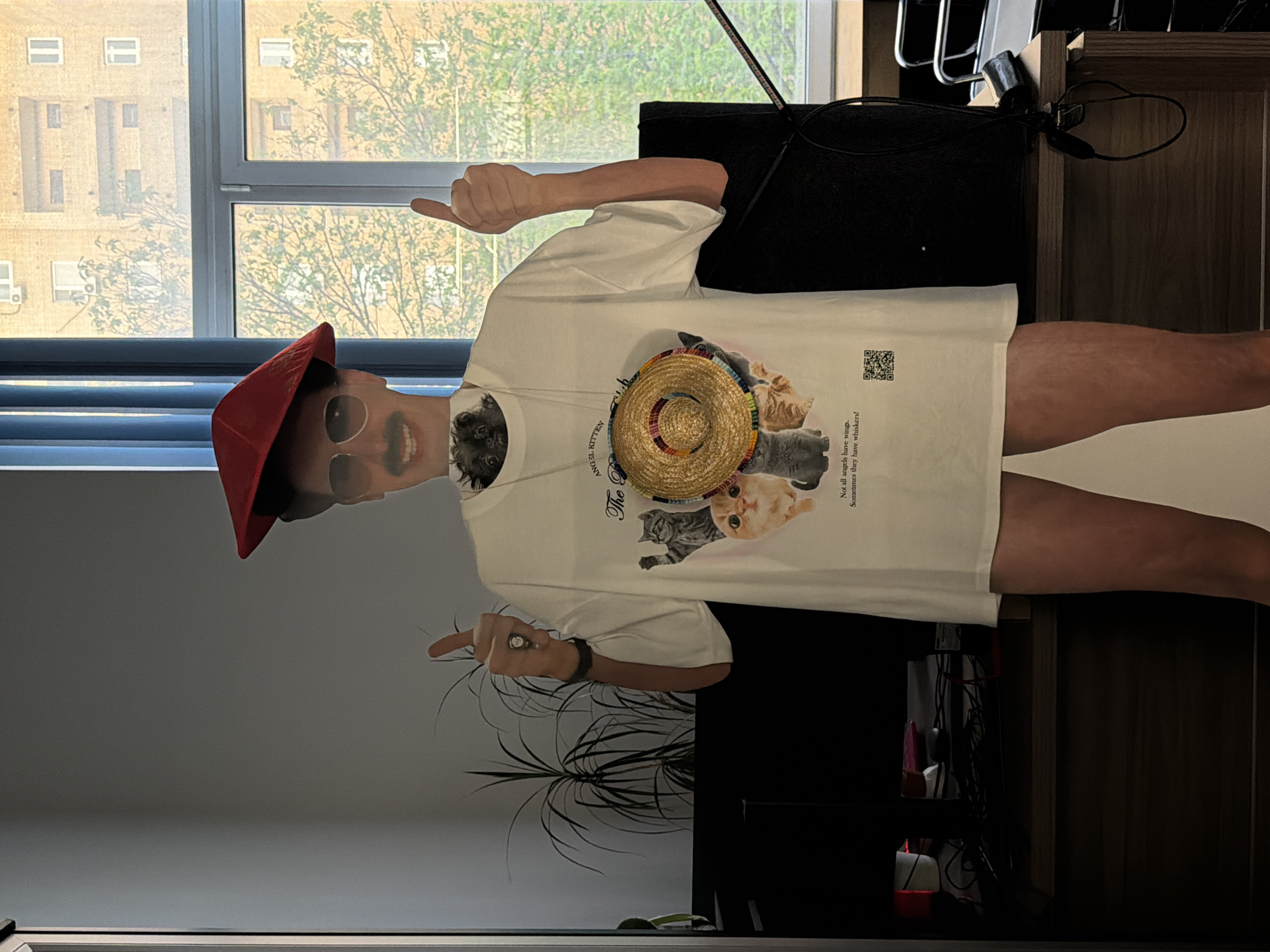 A person is posing indoors. They are wearing a white t-shirt with a cat design and a straw hat on the shirt. The person is also wearing a red hat and sunglasses. They are giving a thumbs up with both hands. A window is visible in the background, showing an apartment building and trees. A plant is partially visible in the lower portion of the picture. The environment appears to be a modern room. The picture was taken in the city of Khimki, Russia.