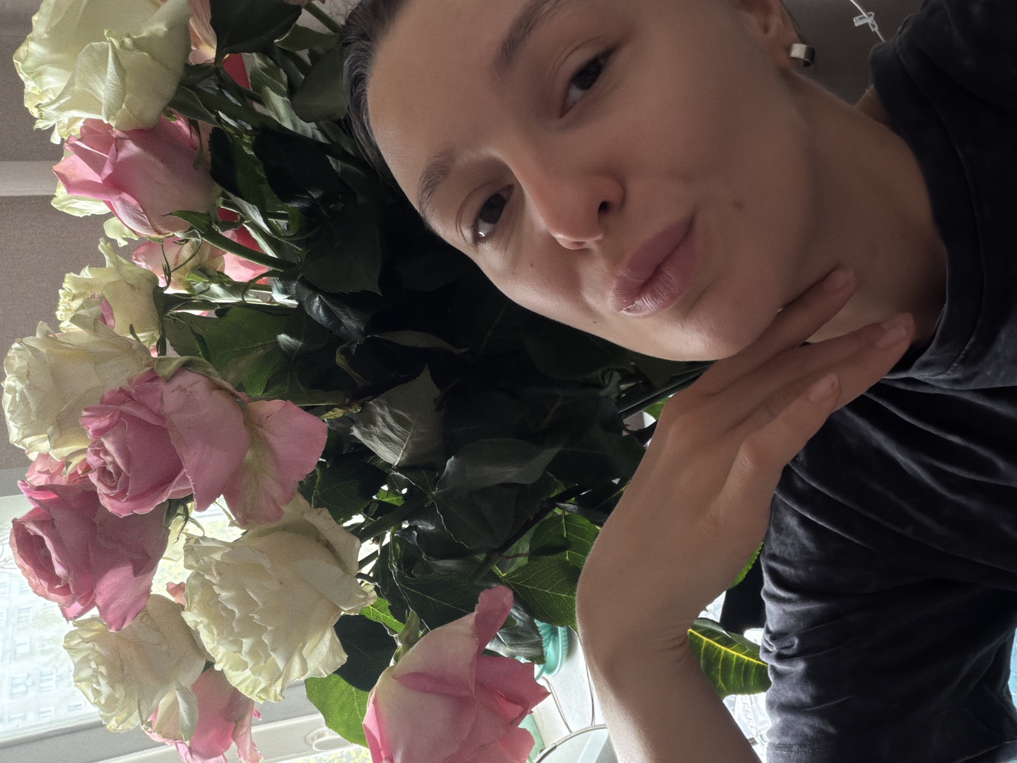 The photograph captures a woman in close proximity to a bouquet of roses. The roses are a mix of white and pink shades. The woman's right hand is positioned near her face. She appears to be wearing a dark-colored top and a small, circular earring is visible. The setting appears to be indoors, with a window in the background. The city and state are unknown.