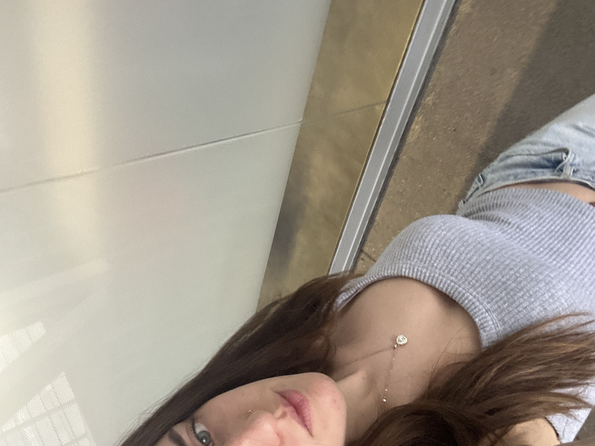A medium close-up, diagonally oriented shot shows a young woman looking directly at the camera. Her head is positioned in the lower left quadrant of the frame, tilted upwards, while her torso extends towards the upper right. She has long, dark brown hair that partially covers her left shoulder and chest. Her eyes are a striking blue-green, and her lips are slightly parted, showing a natural pink hue. A small silver stud pierces her left nostril.
She wears a light grey ribbed tank top or crop top, which reveals a small portion of her midriff where light blue ripped denim shorts or jeans are visible. A delicate gold-toned necklace with a small, circular pendant rests on her collarbone. Another, longer gold-toned necklace with a heart-shaped pendant, possibly adorned with small diamonds, hangs lower on her chest, with a small drop chain visible.
The background consists of a bright, light-colored (possibly white or off-white) wall or partition on the left, with subtle light reflections and faint horizontal lines suggesting panels. To the right of the wall, a vertical metallic strip, appearing gold or bronze, runs alongside a thinner silver metallic frame. Below this frame, a brownish-grey concrete or tiled floor is visible. The overall lighting is soft and even, suggesting an indoor or sheltered outdoor environment during the daytime in Mexico City, Mexico. No visible text is present in the image.