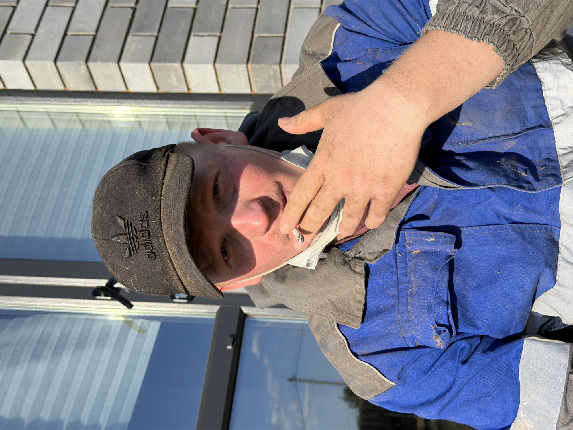 A man is wearing an Adidas cap and a blue and white work outfit, and is holding a cigarette. The location is Chaikovsky, Russia, though the image does not provide specific street details. A building is visible in the background.