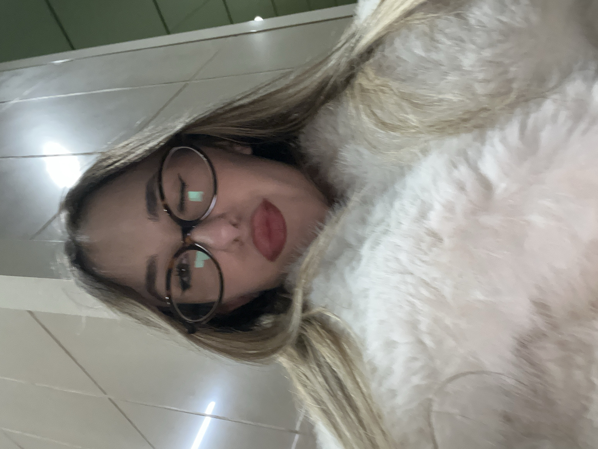 A selfie features a young woman with long, blonde or light brown hair, wearing round-framed glasses. Her face is partially visible, angled towards the camera with her lips slightly puckered in a "duck face" expression. She is clad in a light-colored, possibly fur or faux fur, garment that frames her head. Reflections from overhead artificial lights are visible in her glasses. The background shows light-colored tiled walls and a light green ceiling or panel in the upper left portion of the image, indicating an indoor setting, possibly a public space. The image is rotated 90 degrees clockwise. There is no visible text. The scene is set in Sofia, Bulgaria.