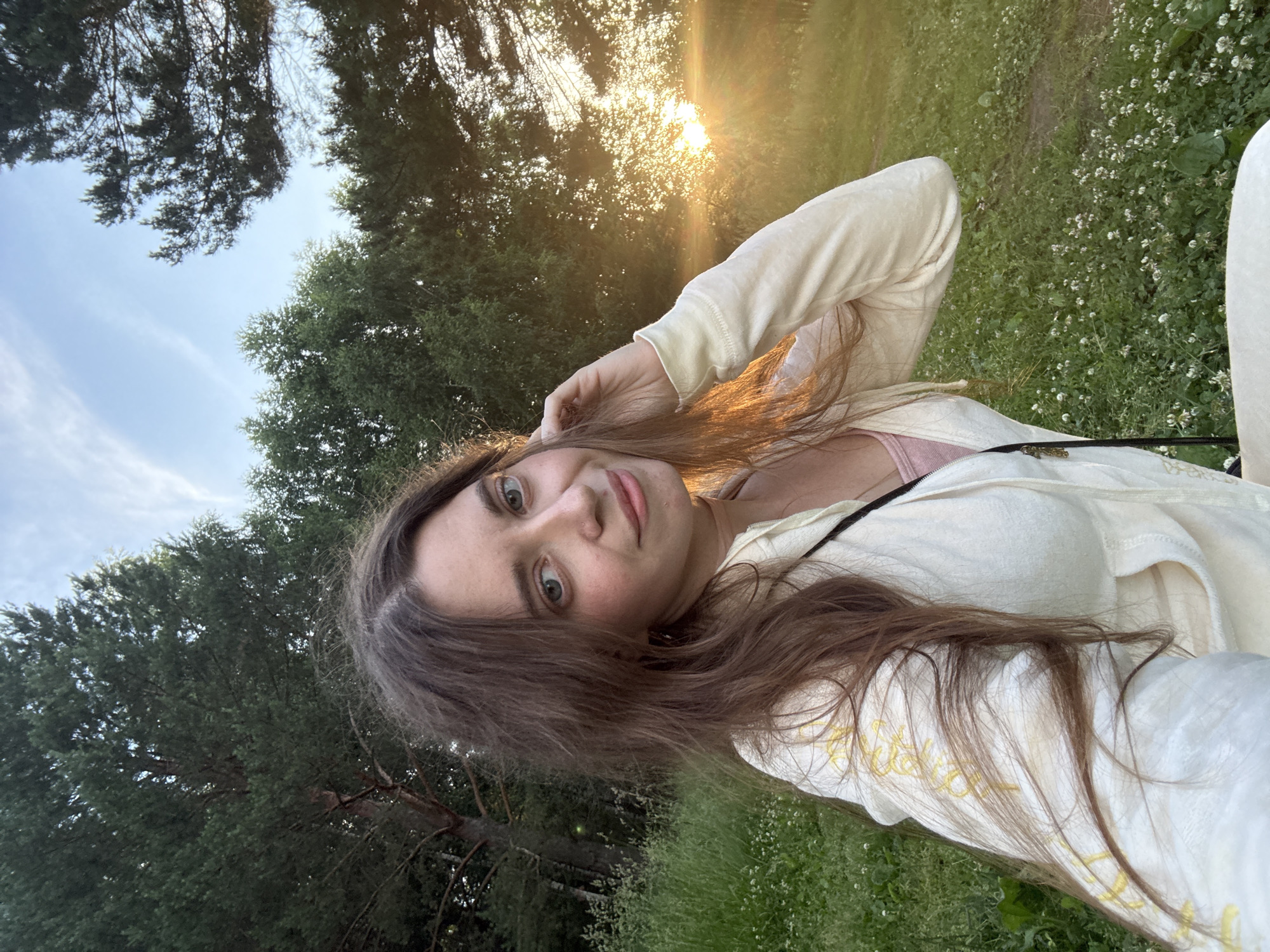 A young woman with long brown hair and light blue eyes is pictured in a selfie taken outdoors at sunset. She is wearing a light-colored hooded jacket over a pink top. The background consists of a grassy field with scattered white flowers, trees, and a sunset with visible sun rays. The woman is touching her hair with her right hand and smiling at the camera. The overall scene suggests a relaxed, outdoor setting during golden hour. The location is unknown, and it is impossible to determine if it is in a city.
