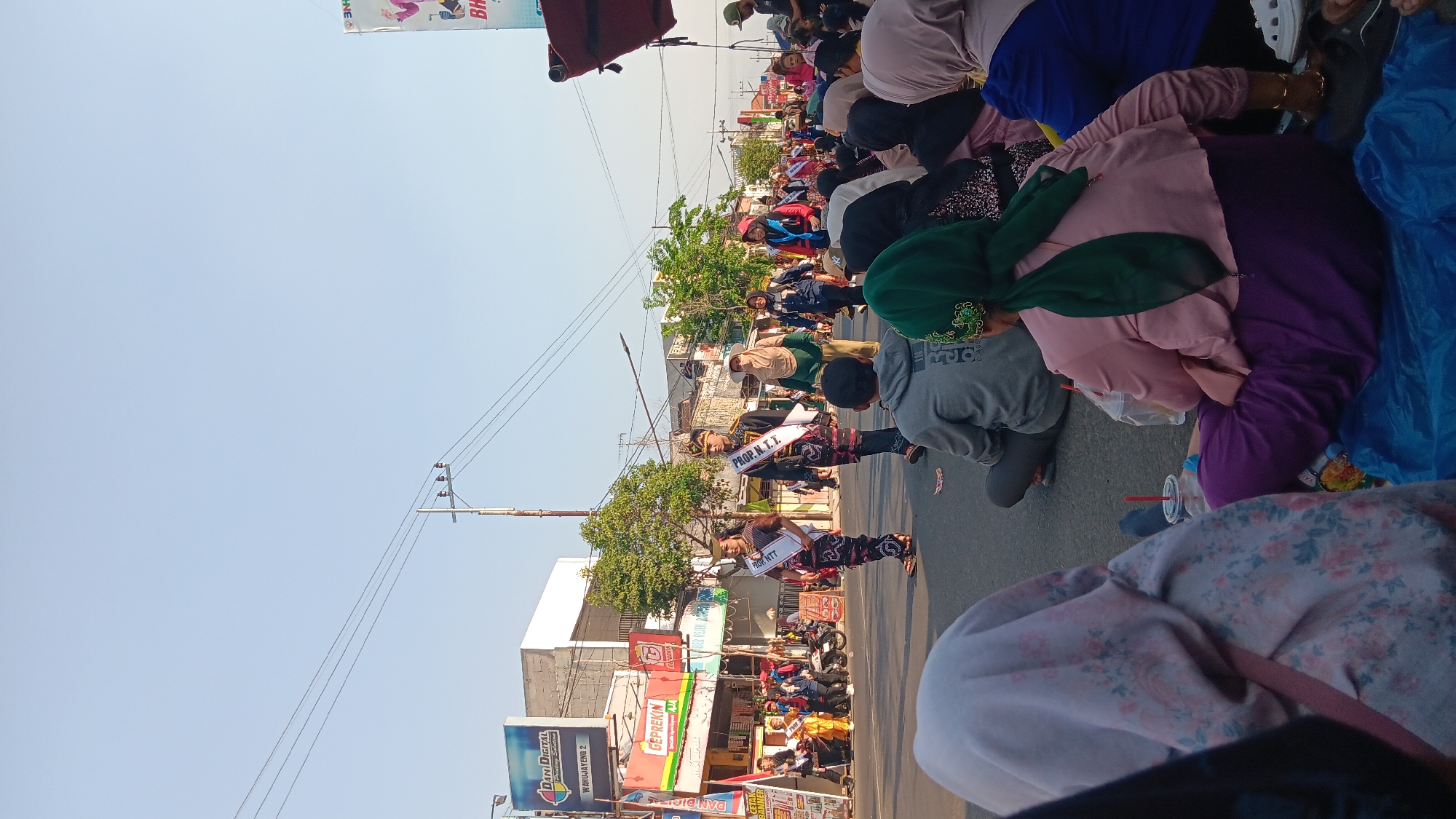 A street parade is in progress in Tanjung Anom, Indonesia. A group of individuals in traditional attire are walking down the street, some wearing sashes that read "PROP. NTT". Spectators are seated on the ground watching the procession. The environment is an urban street with buildings and power lines visible. The sky is clear and blue, indicating daytime.