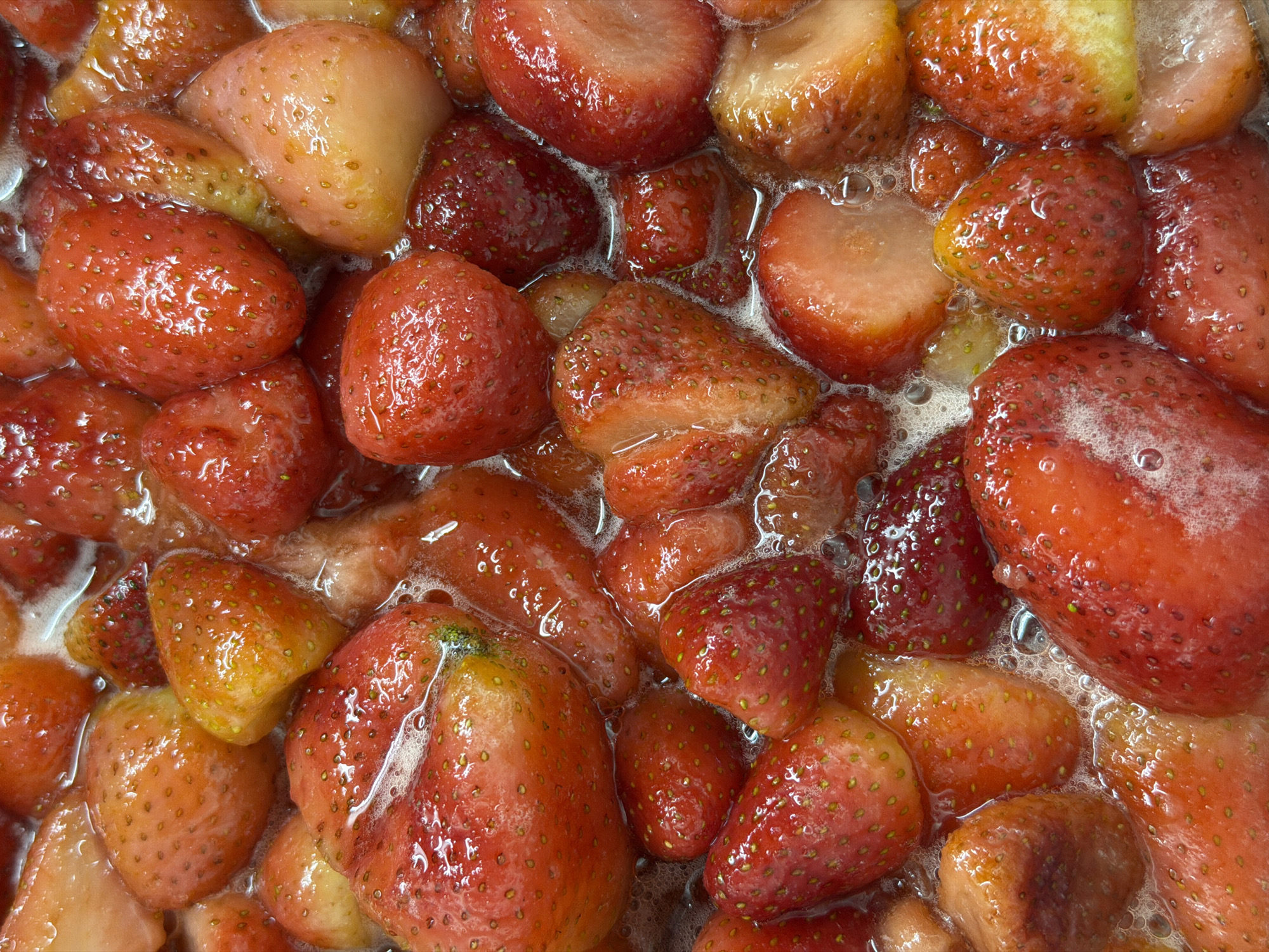 The image shows a close-up of strawberries in liquid. The strawberries vary in color from light orange to deep red. Some are whole, others are cut in half or smaller pieces. The liquid appears slightly viscous, with some bubbles. There is no information to infer that this is from Belgrade, Serbia.