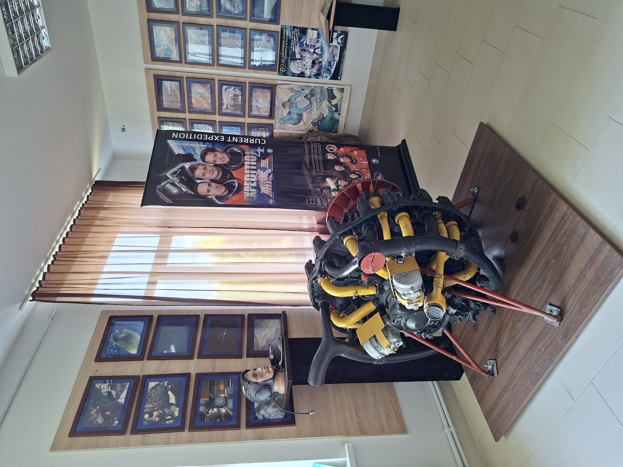 In a room, likely a museum or exhibition space in Ialoveni, Moldova, a large, complex engine component, possibly from an aircraft or spacecraft, is displayed on a wooden platform. The engine is predominantly black and yellow with red support legs. To the left of the engine, a mannequin head wearing a silver helmet with earpieces is positioned on a shelf, accompanied by a small black object.
The walls are adorned with numerous framed photographs, predominantly depicting space-related imagery, including astronaut portraits and celestial scenes. A large vertical banner featuring astronauts and the text "CURRENT EXPEDITION" stands to the right of the mannequin. In the background, a window is covered by sheer brown curtains, allowing diffuse light to enter the room. The floor is tiled with light-colored rectangular tiles.