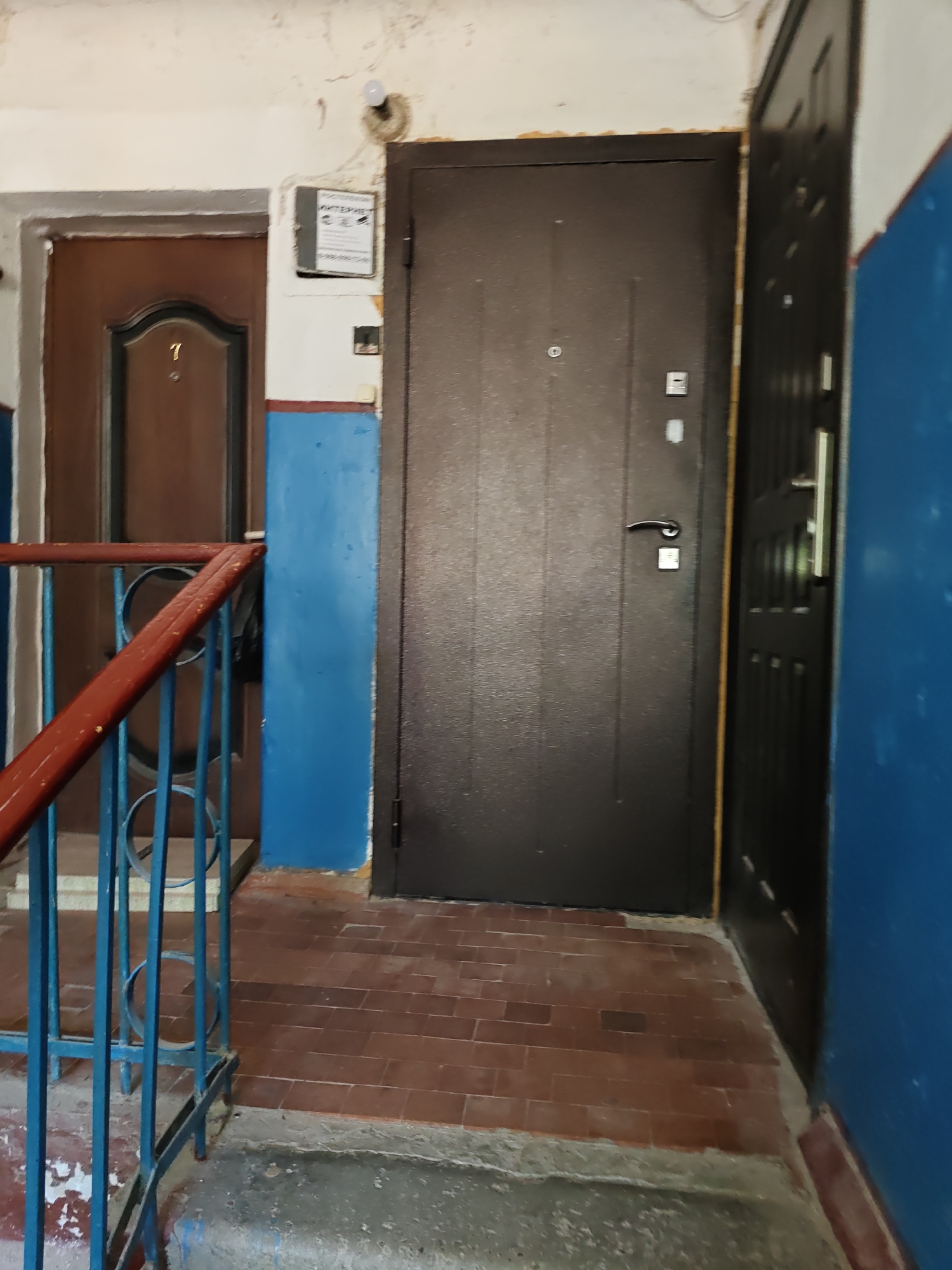 The image shows a hallway with two doors and a stair railing. The door on the left is brown with a number 7 on it and a peephole. The door on the right is dark brown with a silver handle and is slightly ajar. A sign with Cyrillic text and what appears to be security camera icons is mounted on the wall next to the brown door. The walls are painted white and blue, with some peeling paint and cracks. The floor is tiled with reddish-brown tiles. A blue metal stair railing with a red wooden handrail is visible in the foreground. The scene appears to be an interior hallway in an apartment building in Nizhnedonskoy, Russia.