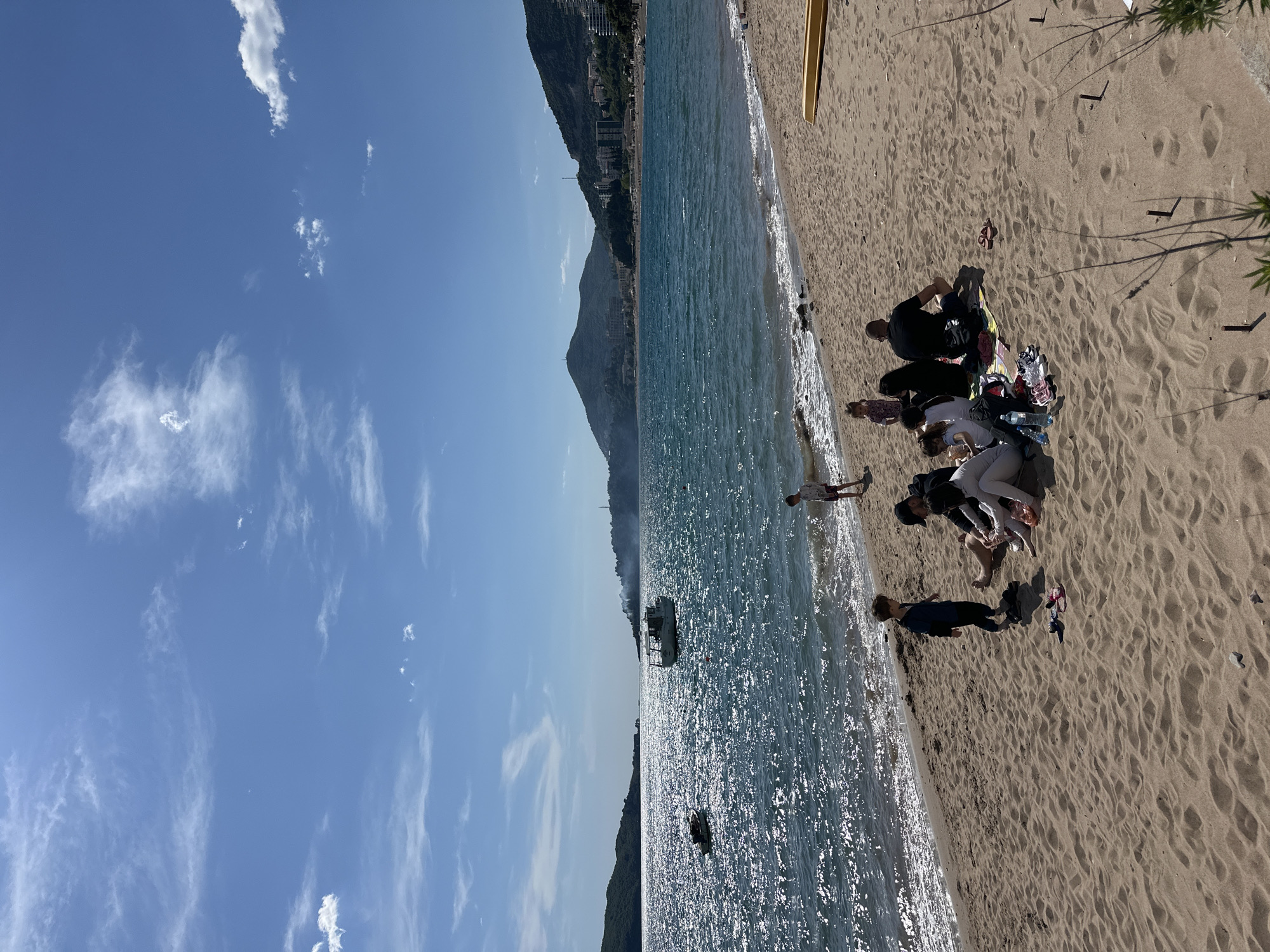 This image captures a vibrant beach scene in Rafailovići, Montenegro, under a bright, sunny sky. The perspective is from the sandy shore, looking out over the sparkling blue-green sea towards a mountainous coastline in the distance.
On the sandy beach, several people are visible. A group of individuals, appearing to be adults and possibly children, are gathered in the mid-ground, sitting or lounging on the sand. They have personal belongings such as bags, clothes, and multiple water bottles scattered around them. Some wear dark attire, while others are in lighter clothing. Further towards the water, a person in dark clothing stands near the lapping waves, and another small child is seen walking towards the water. Footprints and small ripples are noticeable in the sand, suggesting recent activity. Partial views of a yellow kayak or small boat on the sand and the shadows of palm trees or beach umbrellas are visible in the foreground.
The sea, glinting intensely under the bright sunlight, shows gentle waves breaking along the shore. Further out in the water, a white boat is anchored or slowly cruising, and a smaller, dark object, possibly an inflatable or a person, floats closer to the beach.
In the background, steep mountains covered in lush greenery rise from the coastline. Numerous multi-story buildings are interspersed along the mountain slopes, indicating a developed coastal town. The sky above is a clear, vibrant blue with scattered white clouds, ranging from wispy cirrus to more defined cumulus.
The scene depicts a leisurely day at the beach, with people relaxing and enjoying the sunny weather. The strong sunlight indicates it is likely mid-day to early afternoon. There is no visible text in the image.