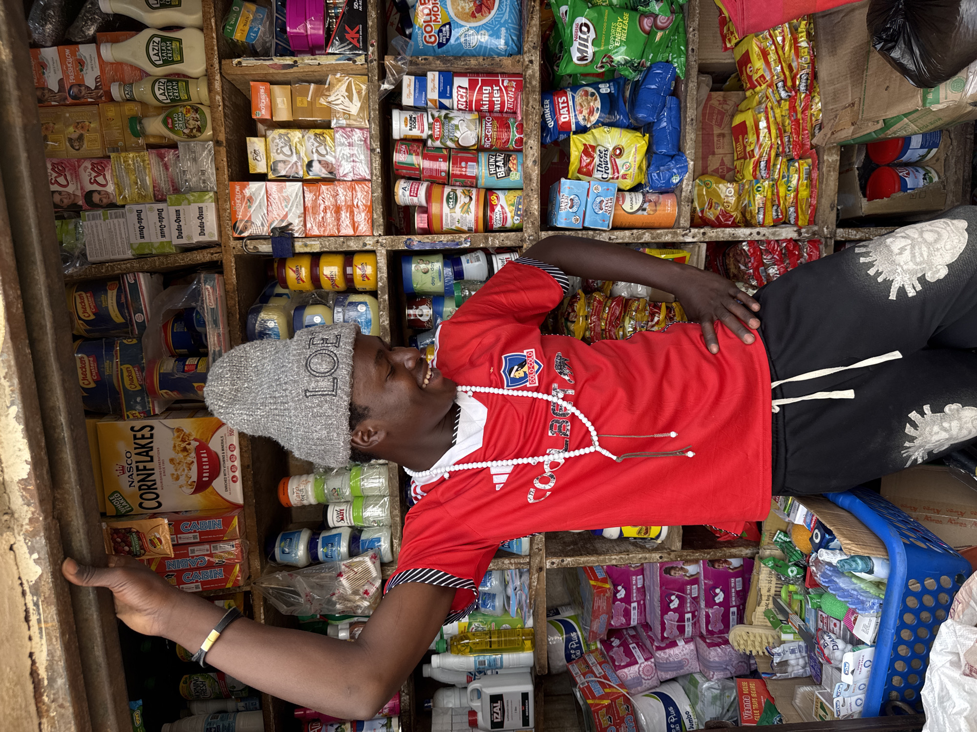 A young Black person wearing a grey beanie and a red shirt with "COLD-COLO" written on it is leaning against a shelf in a small store. They are looking up and smiling. The shelves are packed with various products, including toiletries, food items, and cleaning supplies. Visible text includes brands like "LAZIZ," "Eva," "Dudu-Osun," "NASCO CORNFLAKES," "Checkers Custard," "PASSION," "Nestle MILO," "QUAKER OATS," and "IZAL." The store appears to be a local shop in Jos, Nigeria, filled with everyday consumer goods. The lighting suggests it might be daytime, with artificial lights also present. The person's smile conveys a sense of happiness or amusement.