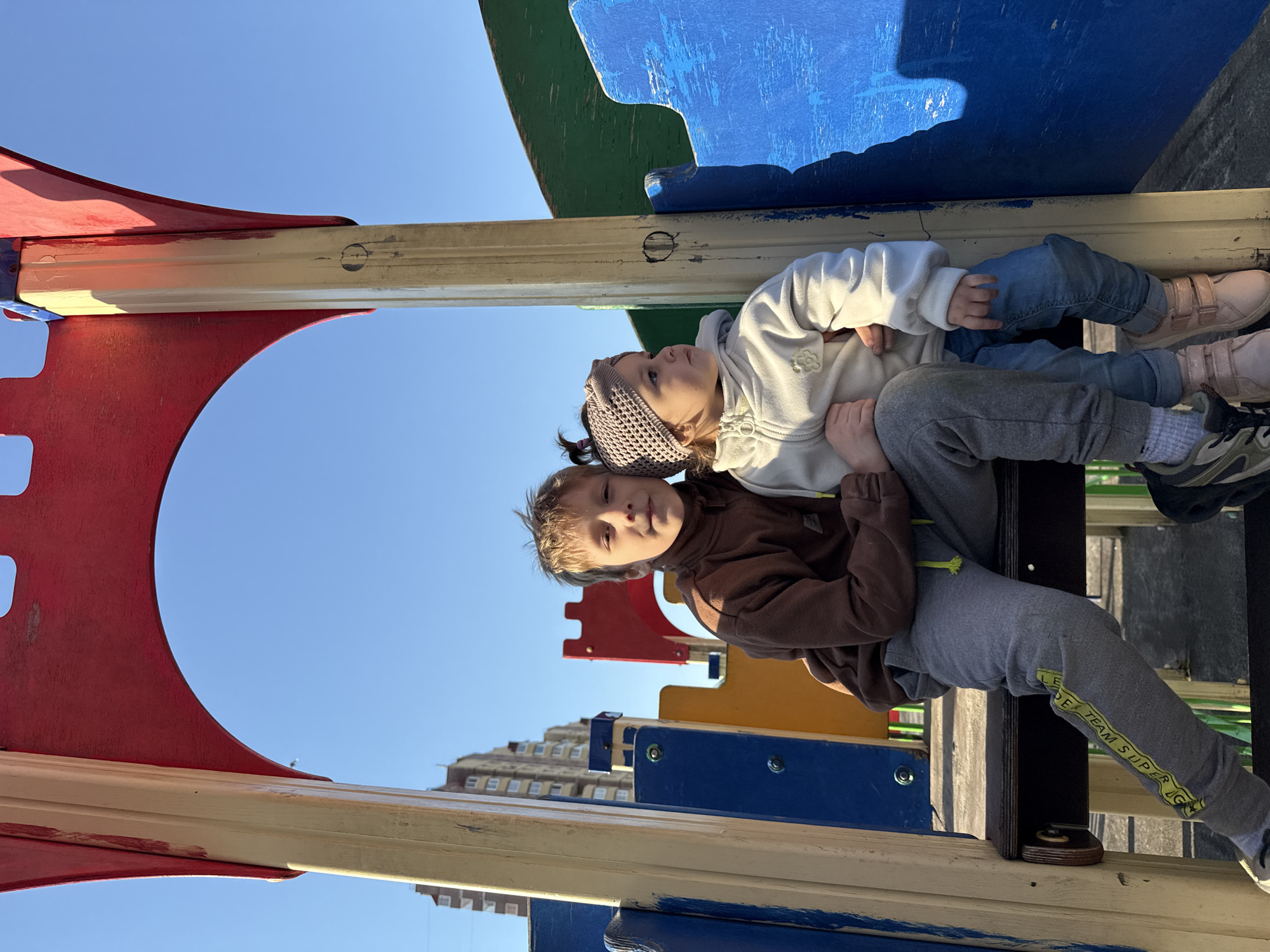 Two children are sitting together on a piece of playground equipment constructed from painted wood and metal. The older child wears a brown jacket and gray sweatpants with yellow text along the leg, and faces forward. The younger child is behind the older child, looking upwards. The younger child wears a white sweatshirt, blue jeans, and a knitted headband. The playground equipment has elements of red, yellow, blue, and green paint. In the background is a clear blue sky and a multi-story building. Based on the architecture and surrounding details, it is plausible this image was taken in Tobolsk, Russia.