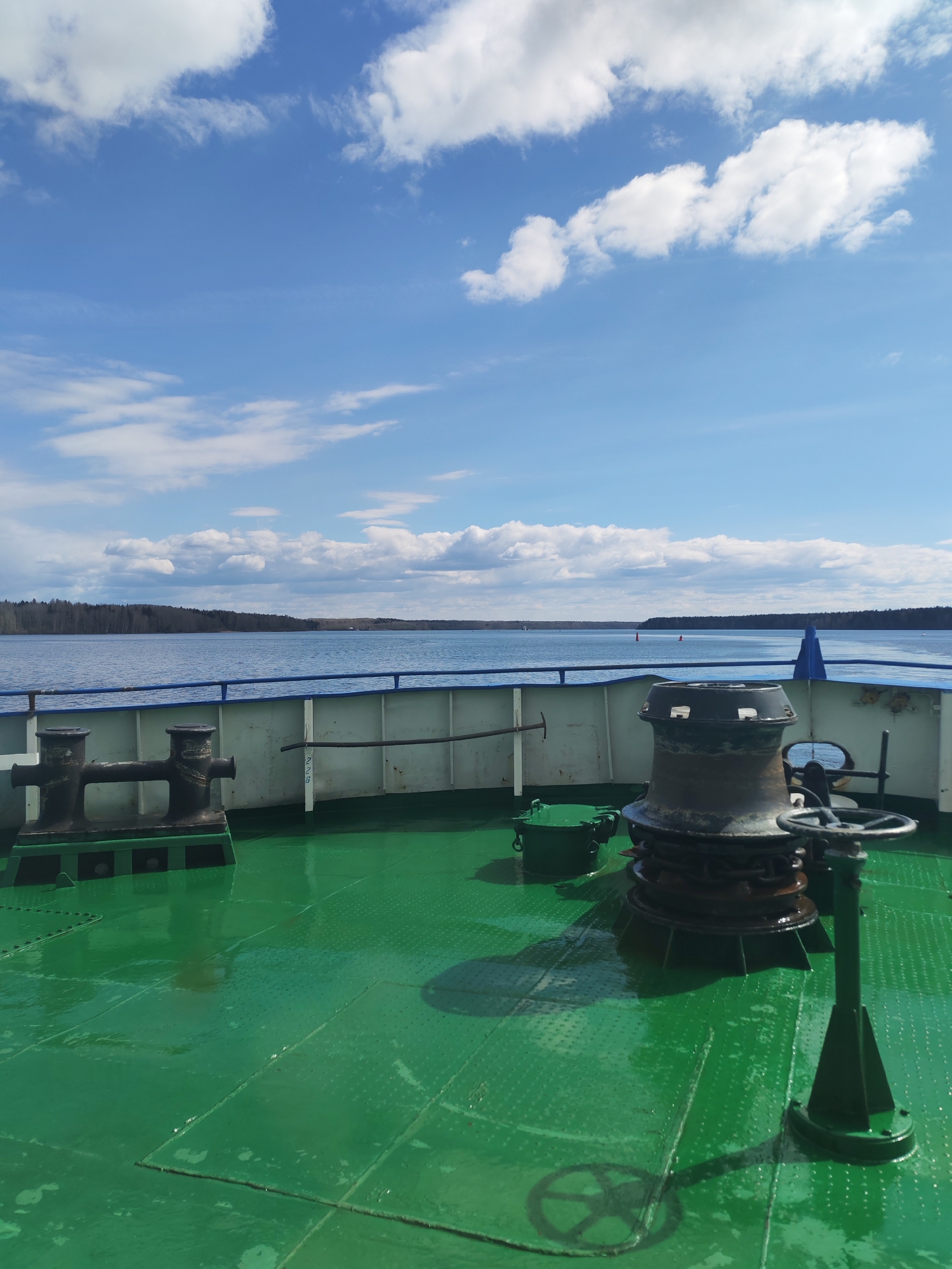 The image captures a sunny daytime view from the front deck of a vessel, looking out over a wide body of water towards a distant forested shoreline. The foreground is dominated by the ship's green, textured non-slip deck, which appears wet in places, possibly from recent rain or splashes.
On the left side of the deck, heavy black metal bollards are firmly bolted down. Towards the right, a large, cylindrical, dark metal capstan or windlass with a chain wrapped around its base is prominent, alongside a small, round green container or hatch. Further to the right, a dark green pedestal topped with a steering wheel-like handle stands, casting a clear shadow on the wet deck.
A blue-topped railing with a white lower section separates the deck from the calm, blue water. Faint black text or numbers, "226," are visible on the white part of the railing. Beyond the railing, the expansive water surface is tranquil, reflecting the clear sky. In the mid-distance, a small red buoy is visible on the water.
The background features a continuous dark green, forested shoreline stretching across the horizon, suggesting a large lake or a wide river. Above the land and water, the sky is a vibrant blue, adorned with scattered, fluffy white cumulus clouds, indicating fair weather. The sun is high, casting distinct shadows and brightly illuminating the scene. The overall impression is one of peaceful travel on a clear day.