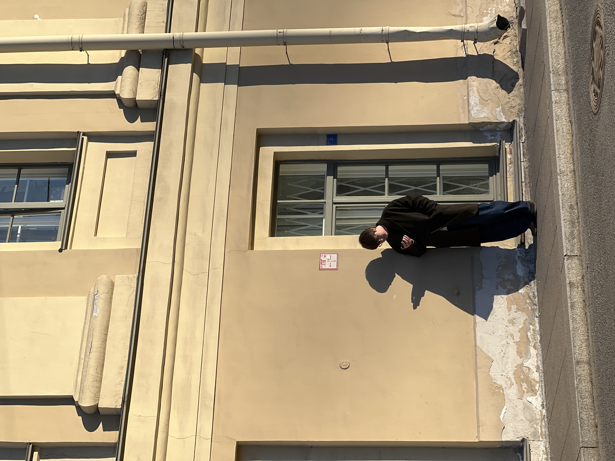 A man in a black coat and blue pants leans out of a window on a yellow building. The building's facade has architectural details, including a horizontal pipe and vertical columns. A sign with red and white markings is visible near the window. The building's paint is peeling near the window, and a sidewalk runs along the building's edge. The photo suggests the scene is in Saint Petersburg, Russia, based on the architectural style.
