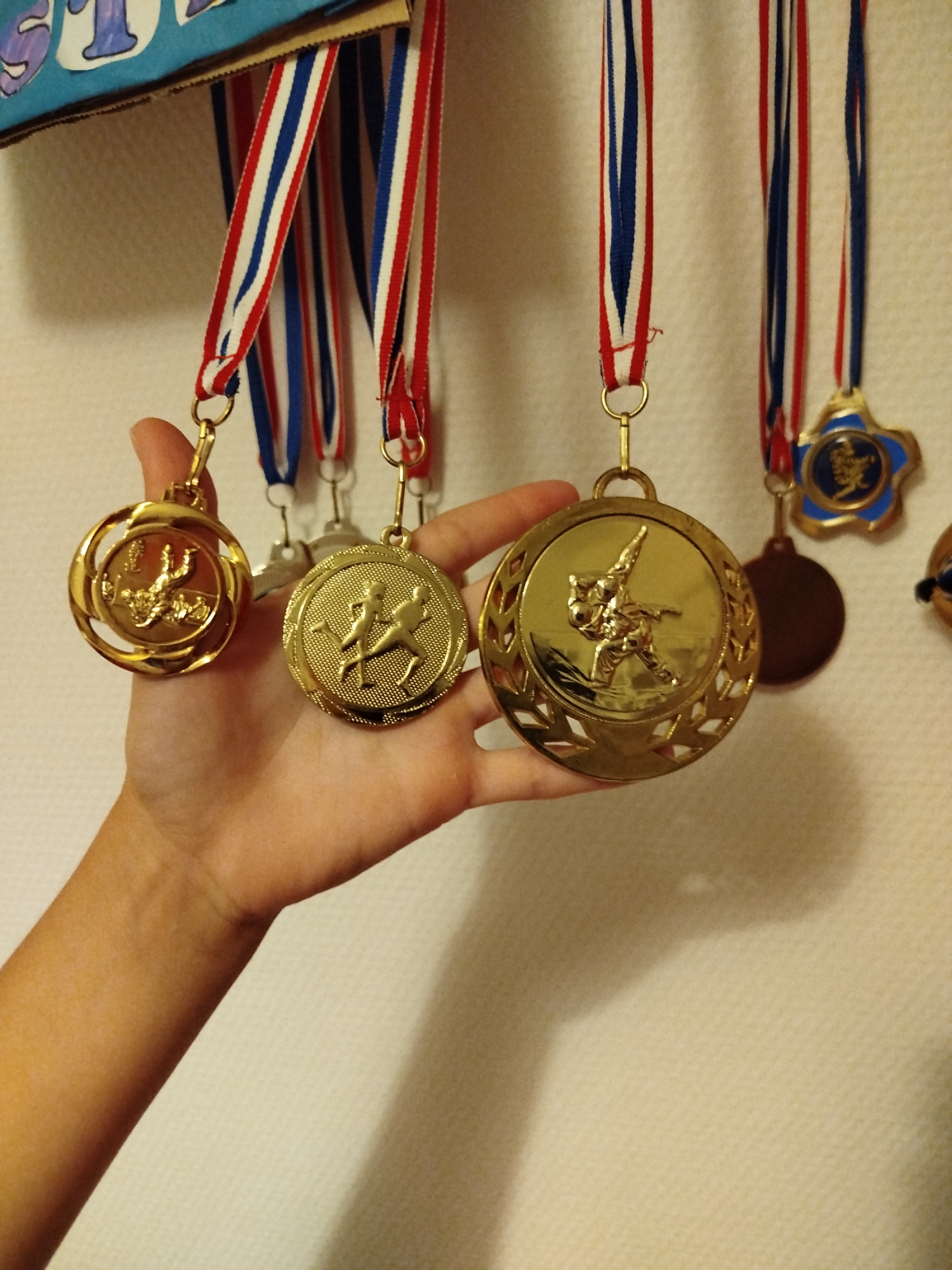 The image shows a hand holding up three gold medals, each attached to a red, white, and blue ribbon. The medals are hanging from a cardboard display that has some lettering on it, but it is mostly obscured. The medals themselves feature embossed figures of athletes. One medal shows a swimmer, another shows two runners, and the third shows a person in what appears to be a martial arts stance. In the background, there is a textured wall. The image does not contain any elements that would allow for the determination of a specific location within the city of Saint-Claude, France.
