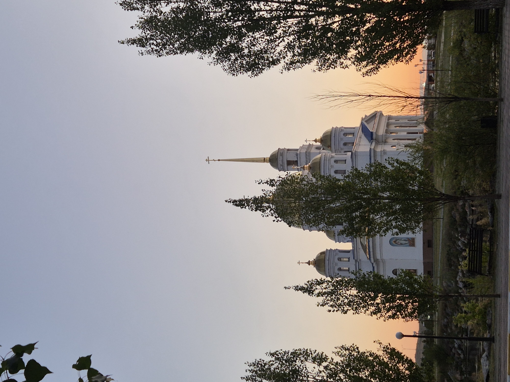 The image shows a large, white, domed cathedral with golden cupolas, possibly an Orthodox church, located in Kokshetau, Kazakhstan. The cathedral is the central focus of the image, with its classical architectural style, featuring columns and a tall spire topped with a cross. Several trees with green foliage frame the cathedral, with some in the foreground obscuring parts of the structure. The sky exhibits a gradient of soft blue at the top transitioning to a warm orange and pink sunset hue near the horizon. In the lower part of the image, there are hints of a park-like setting, with a paved walkway, a bench, and a lamppost. The overall scene suggests a peaceful, serene evening setting in Kokshetau.
