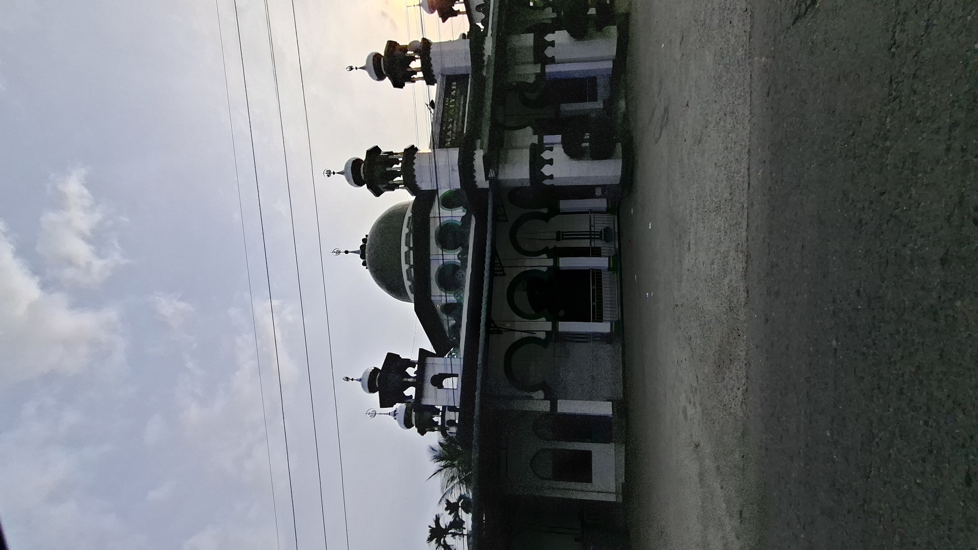 The image shows a mosque in Paya Bengkuang, Indonesia. The mosque has a large green dome and several smaller domes, as well as minarets. The mosque is white and green with arched windows and doorways. There are power lines in front of the mosque. The ground in front of the mosque is paved with asphalt and gravel. The sky is cloudy. The image appears to have been taken at dusk.