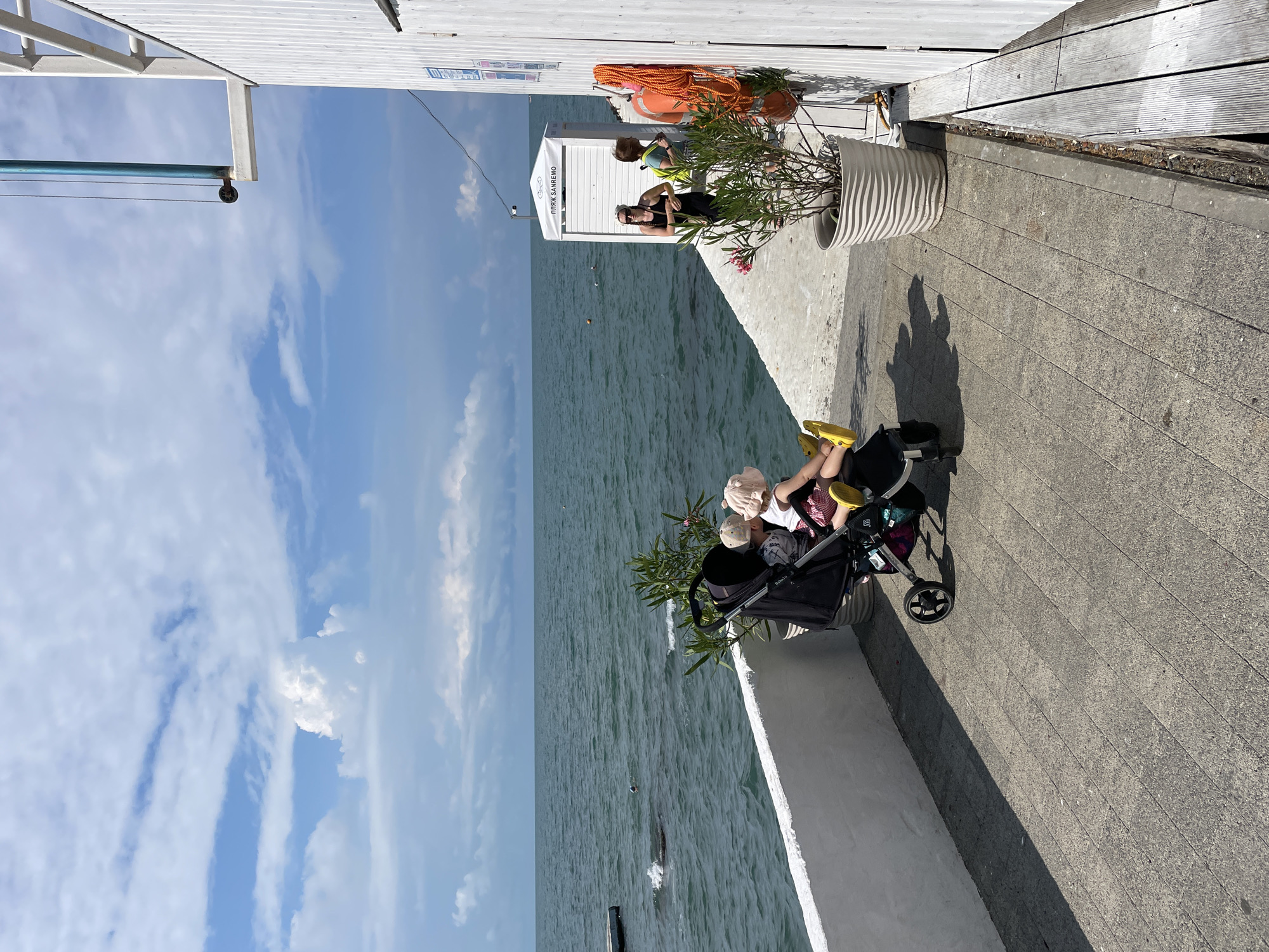 In Sochi, Russia, on a sunny day, two children sit in a stroller facing the sea. The sea is a deep blue with whitecaps. A white building with a green awning is to the right of the frame, and a life preserver hangs on the side of the building. A small white hut with the words "пляж SANREMO" (Beach Sanremo) written on it is behind the stroller. The ground is paved with grey tiles. A plant in a pot sits to the right of the stroller. Two adults stand in the background near the white hut.