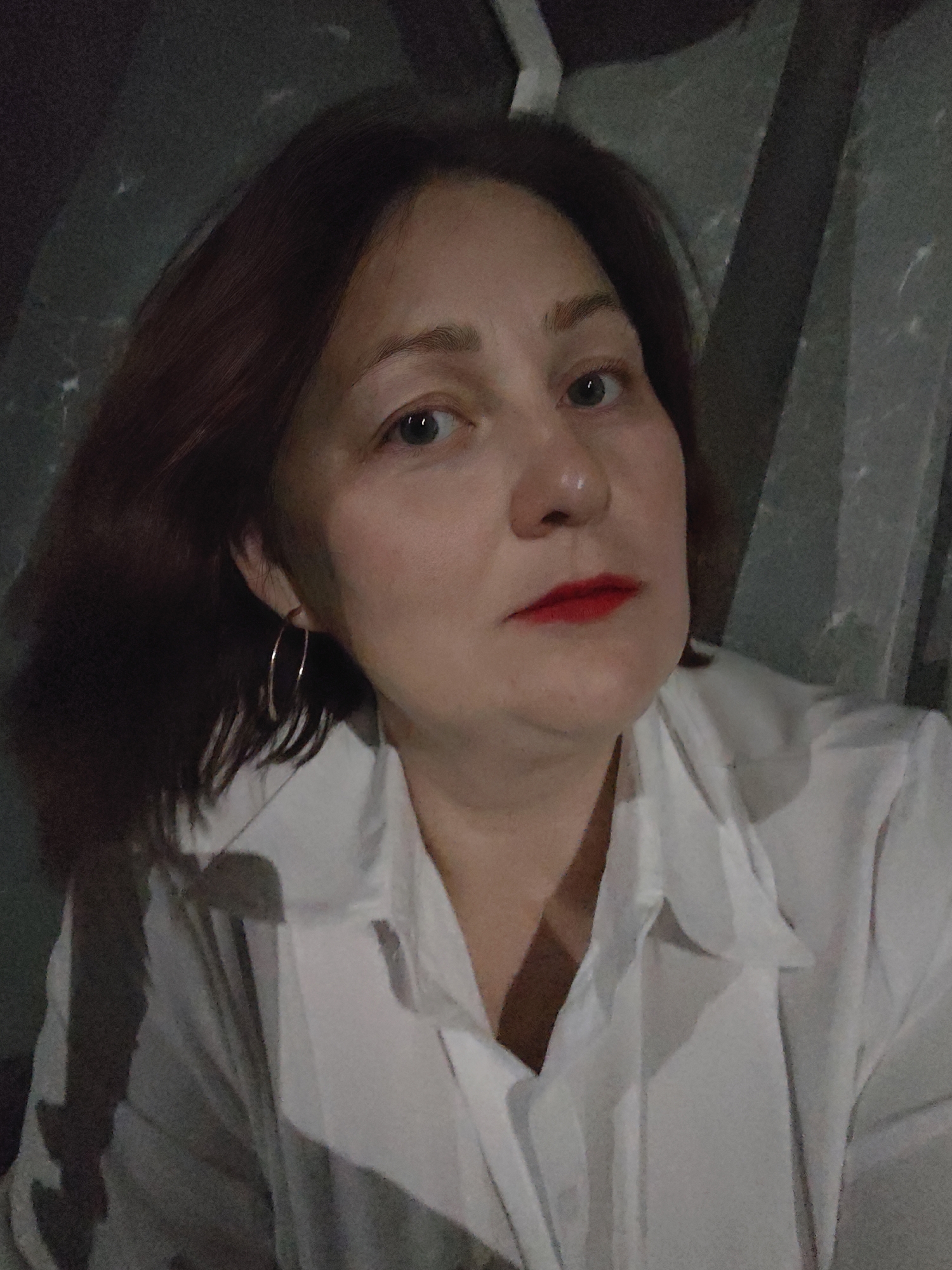 A middle-aged woman with auburn hair and red lipstick is centered in the frame. She is wearing a white, button-down shirt and hoop earrings. The background is poorly lit and appears to be a wall with gray, angular shapes. The photo was likely taken indoors with low lighting conditions. The location mentioned in the prompt (Markovskii, Russia) cannot be verified from the image.