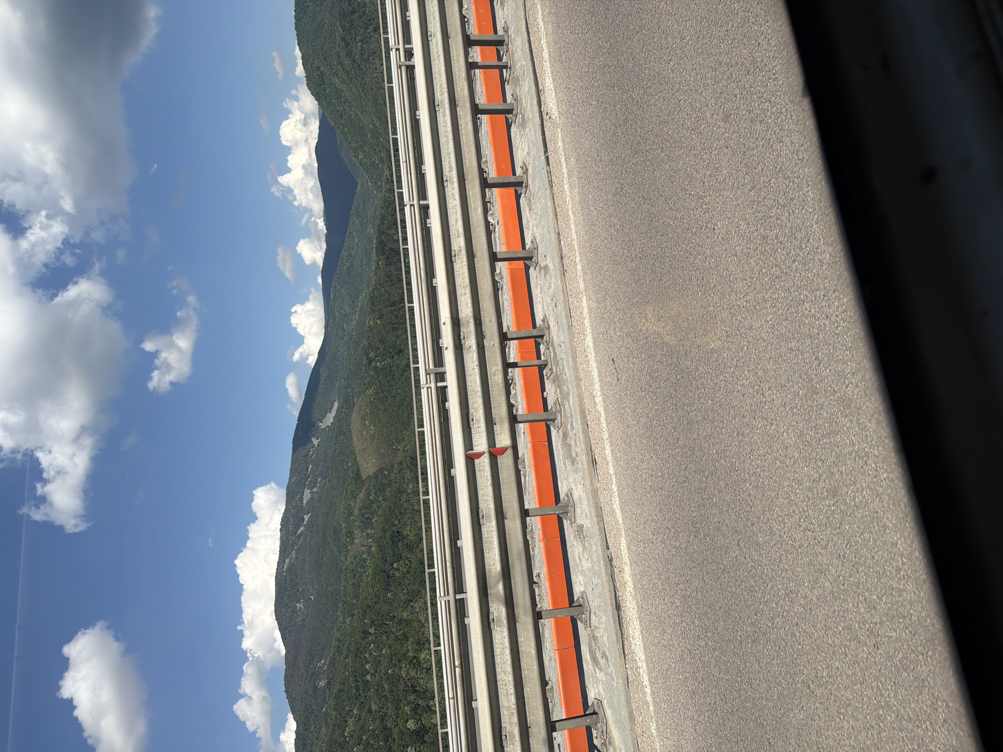 The image shows a view from a bridge with a mountainous landscape in the background. The bridge has a metal guardrail with orange accents. The sky is blue with white clouds. The mountains are covered in green trees, and there are some bare patches on the slopes. The foreground shows the asphalt road of the bridge. No people are visible in the image. This scene is consistent with the location of the Vrachanski Pass bridge, which is located near the town of Vrachesh, Bulgaria, and offers views of the Balkan Mountains.