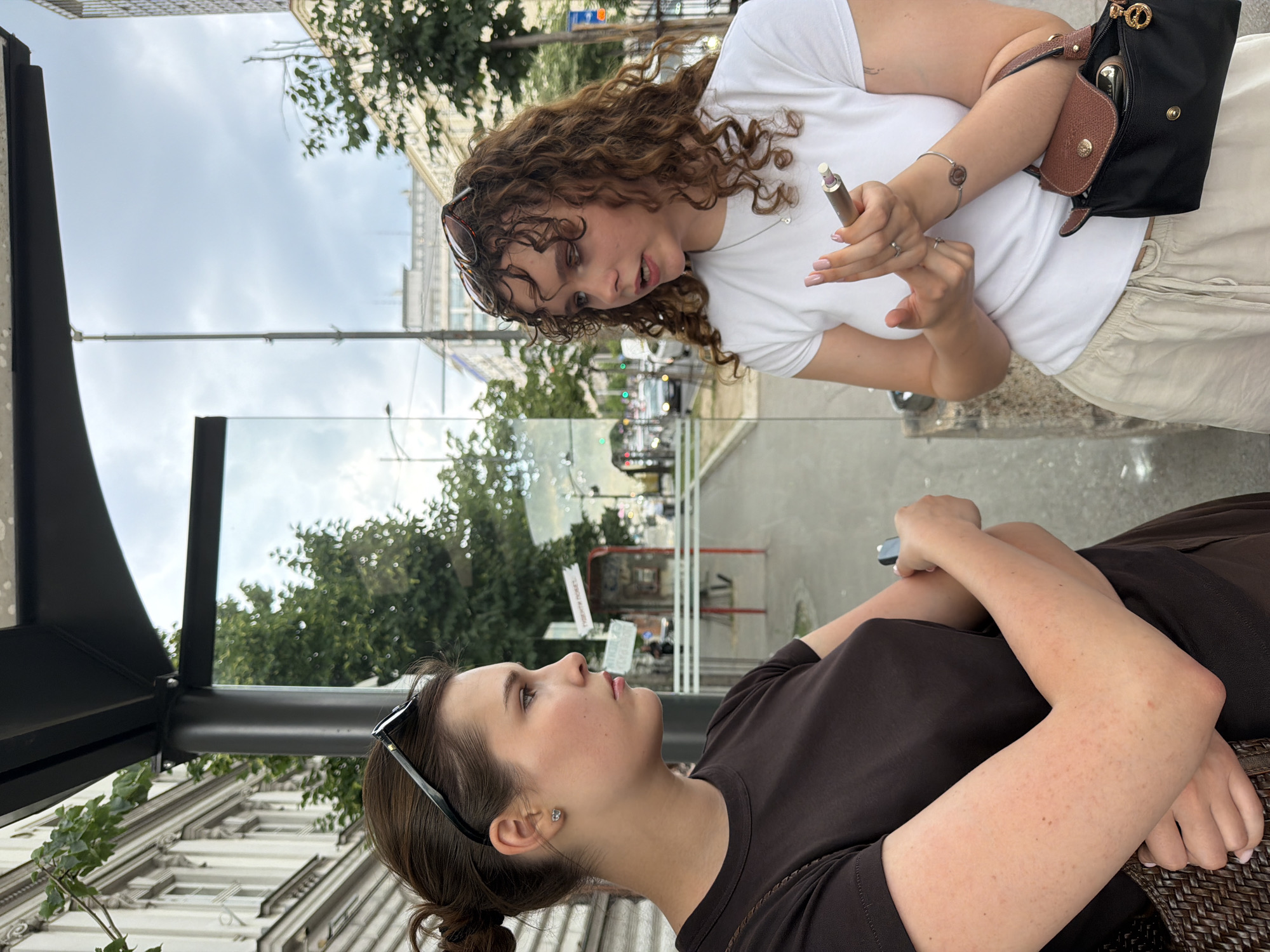 Two women are seated outdoors at a bus stop in Belgrade, Serbia. One woman is seated wearing a brown top, with sunglasses on her head and her arms crossed. She is looking upwards at the other woman. The second woman is standing, leaning in slightly towards the seated woman. She is holding a cosmetic tool in her right hand. She is wearing a white shirt, light-colored pants, a purse, and has curly hair and sunglasses on her head. The bus stop has a glass panel and a black frame, and a woven bench. Through the glass, a street scene with trees, cars, and buildings is visible. The buildings are light-colored with classical architectural features. The sky is overcast.