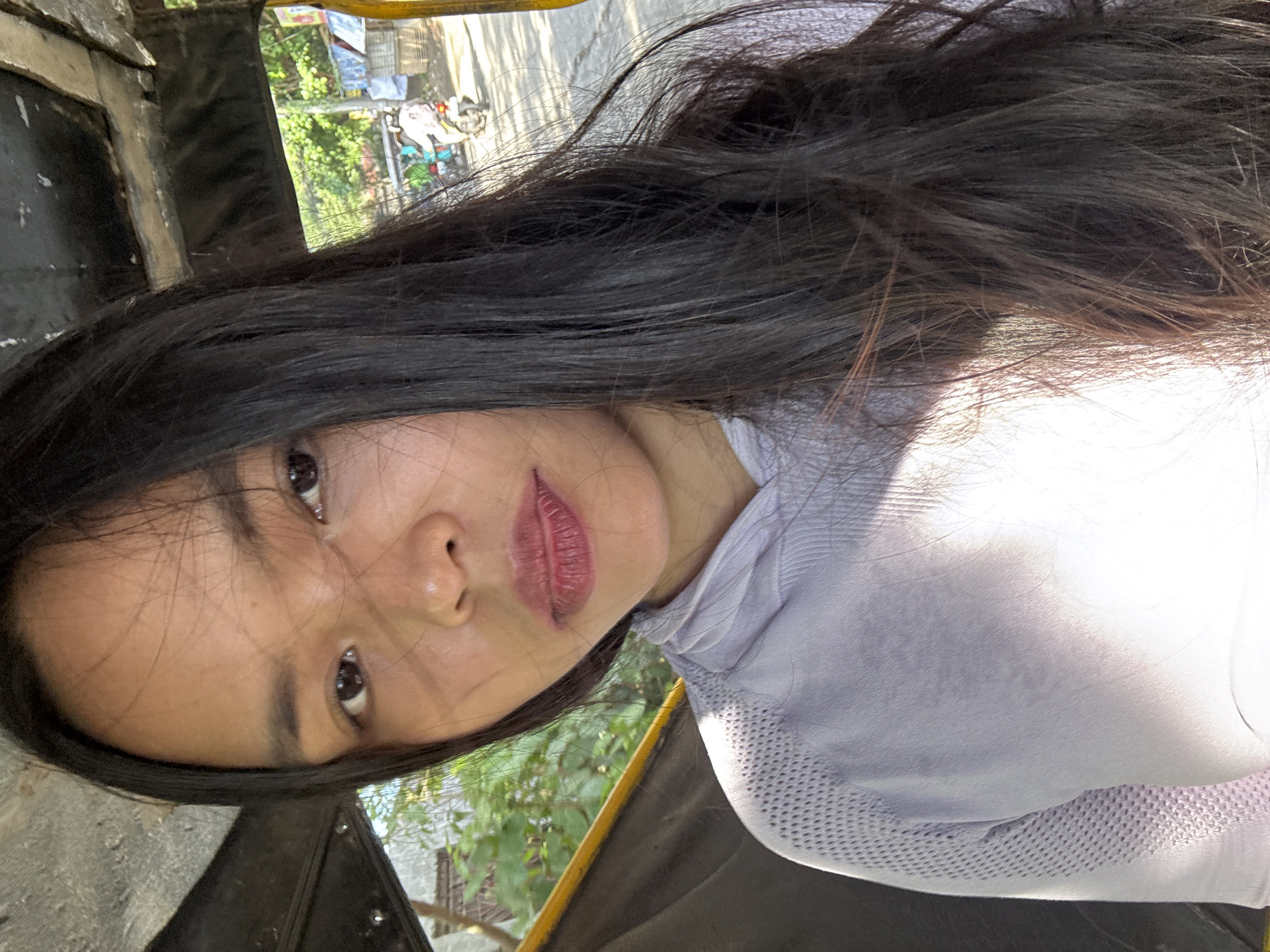 A young woman with long, dark hair is taking a selfie inside what appears to be a tricycle. The vehicle's dark interior, with yellow trim visible, suggests it's likely operating on a street in Davao City, Philippines. Sunlight filters through, casting shadows on her light-colored shirt. In the background, a bustling street scene unfolds with greenery, houses, and a motorcycle carrying a passenger, further reinforcing the urban setting. The woman's gaze is directed at the camera, with a neutral expression.