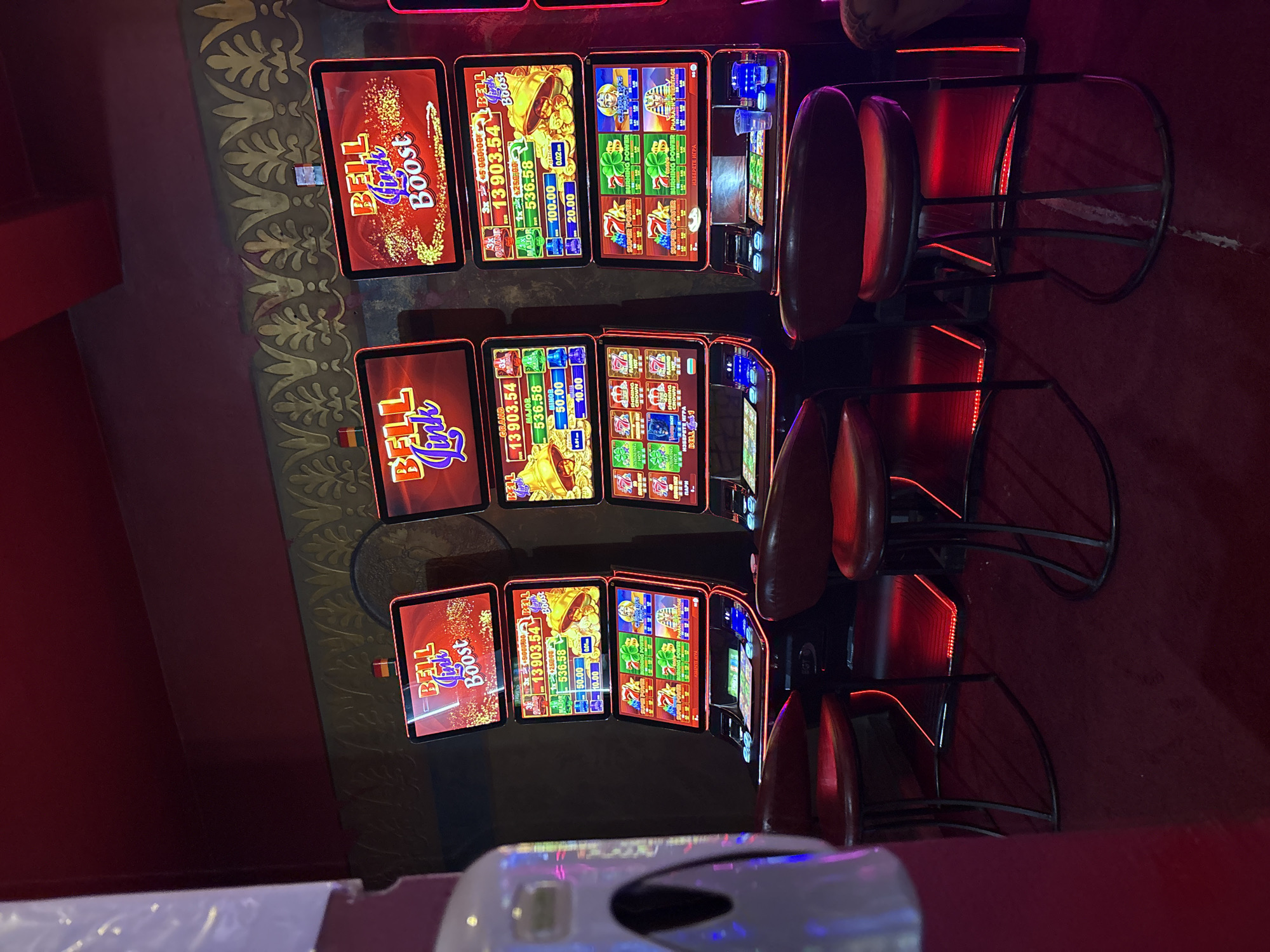 The image shows a close-up, slightly angled view of several slot machines in a dimly lit casino, likely in Sopot, Bulgaria. The machines display vibrant graphics and text related to gaming, including "BELL LINK BOOST," "GRAND," "MAJOR," and "MINOR," along with numerical values indicating jackpots or credits. One machine also shows symbols like pharaohs, clover leaves, and lucky sevens.
In the foreground, part of a white device with a dark circular opening is visible, possibly a hand sanitizer dispenser or a similar amenity. In the background, the red upholstered seats of gaming chairs are partially visible, as is the ornate, gold-patterned wallpaper that lines the wall. The lighting is predominantly red, emanating from the machines and possibly from ambient casino lighting, creating a warm and somewhat moody atmosphere. The overall scene suggests a typical casino environment, likely in the evening or at night, where people are engaged in playing the slot machines. There are no people clearly visible in the image, but the presence of the chairs implies recent or ongoing activity. The text on the machines indicates the games are in progress, with current jackpot amounts displayed.