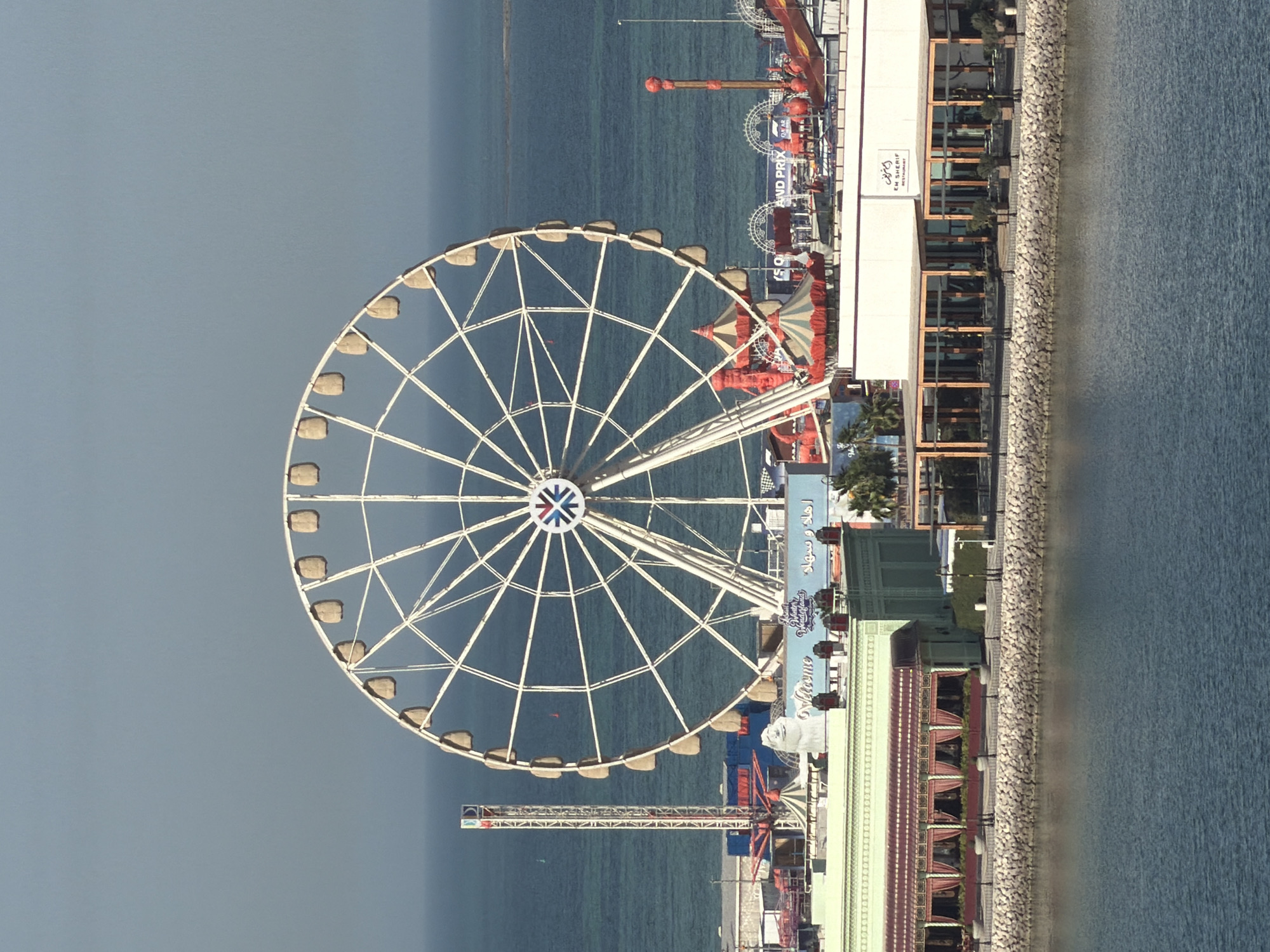 This detailed image captures an amusement park and waterfront area set against a vast body of water. A large white Ferris wheel, featuring light-colored gondolas and a multi-colored hub logo, dominates the central background. To the left of the Ferris wheel, a tall drop tower ride stands prominently. Below and around the Ferris wheel are various amusement park structures, including red and white striped carnival tents, additional red-colored rides, and multiple signs. Notable signs display "Welcome," "Winter Wonderland," "GRAND PRIX," and Arabic script, along with a large white sculpture of a face (possibly a snowman) near the "Welcome" sign.
In the mid-ground, a series of buildings line a rocky waterfront. On the left, a building with a green roof and a maroon awning covering its windows is visible. To its right, another building with a white facade features an outdoor seating area under a wooden pergola-style roof. A sign on this building clearly reads "EM SHERIF RESTAURANT" in English and Arabic. Scattered palm trees are visible around these buildings.
The foreground is dominated by a calm, dark blue-green body of water, likely the sea or a large bay, separated from the land by a grey rocky breakwater. The water extends to the horizon, where a faint landmass or small island is discernible on the right. The sky is a clear, light blue-grey, suggesting fair weather. No people are distinctly visible, and the scene appears static.