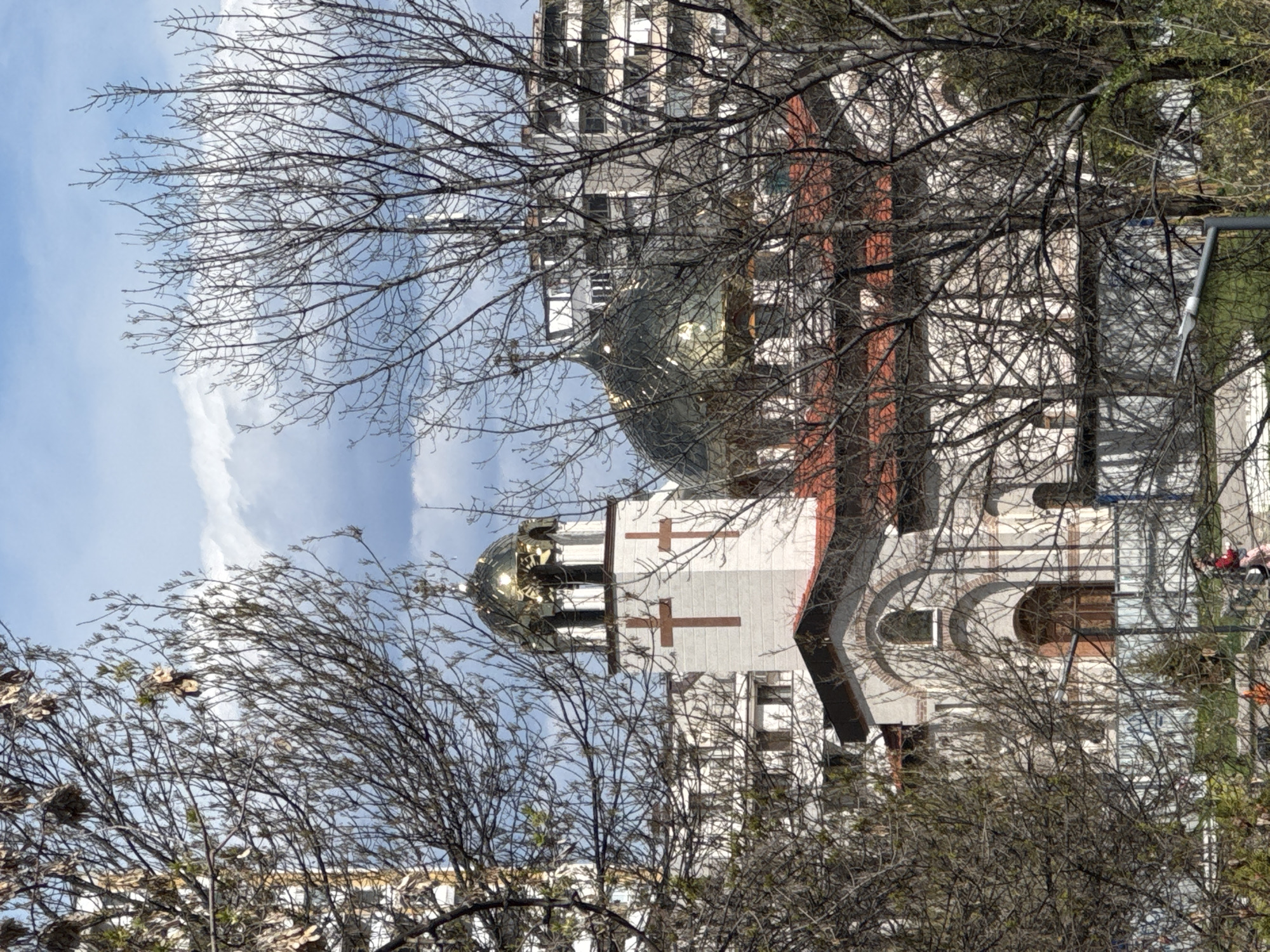The image shows a church with a golden dome in Sofia, Bulgaria, viewed through bare tree branches. The church has a white facade with brown crosses and a red roof. A taller building is visible in the background. The sky is blue with white clouds. The foreground is filled with bare tree branches.