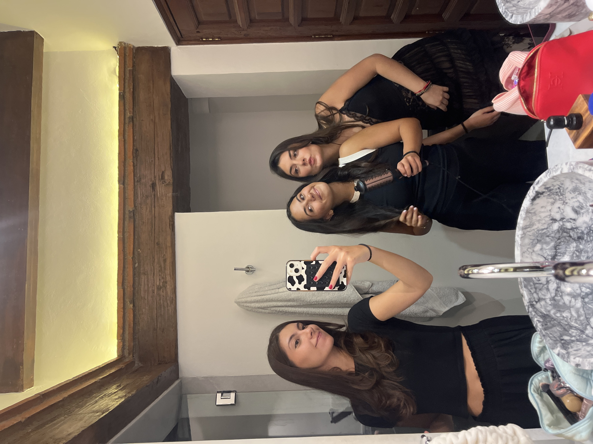 In a well-lit bathroom, three young women are taking a mirror selfie. The woman on the left, holding the phone, is wearing a black crop top and black pants. She is smiling and looking at the camera. The woman in the middle, to her right, is wearing a black sleeveless top and black pants. She is holding a curling iron and looking at the camera. The woman on the right is wearing a black lace dress and is also looking at the camera. The bathroom features exposed wooden beams on the ceiling, a marble sink, and a wooden door. The scene appears to be in a residential setting, possibly a home or a hotel. The presence of a makeup bag with brushes and other toiletries suggests the women might be getting ready for an event. The overall ambiance is casual and fun.