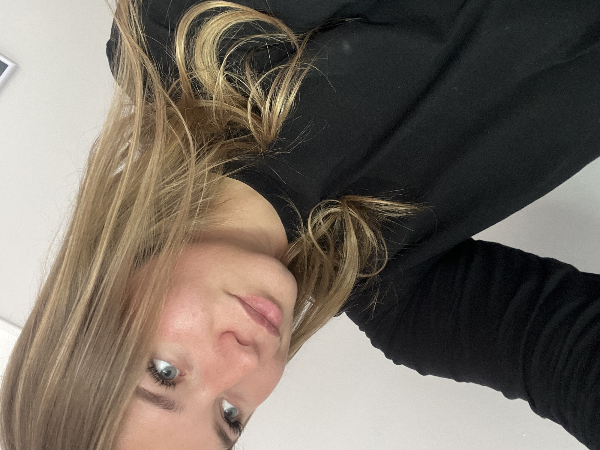 The image is a portrait of a young woman, likely taken indoors in Budva, Montenegro. The photo is rotated 90 degrees counter-clockwise, with her head tilted and her face oriented towards the viewer from a low angle. She has long, wavy hair in shades of blonde and light brown, which frames her face and cascades over her shoulders and a black top. Her eyes are light blue-grey, and she maintains a direct gaze with a neutral, slightly pensive expression. Her skin appears fair.
She is wearing a simple black long-sleeved top. The background is a plain, light-colored wall (likely white or off-white), suggesting an uncluttered indoor setting. No other objects or text are visible in the frame. The lighting is soft and even, characteristic of an indoor environment, making the time of day and external weather conditions indiscernible. The primary activity is the woman posing for the camera, possibly taking a selfie.