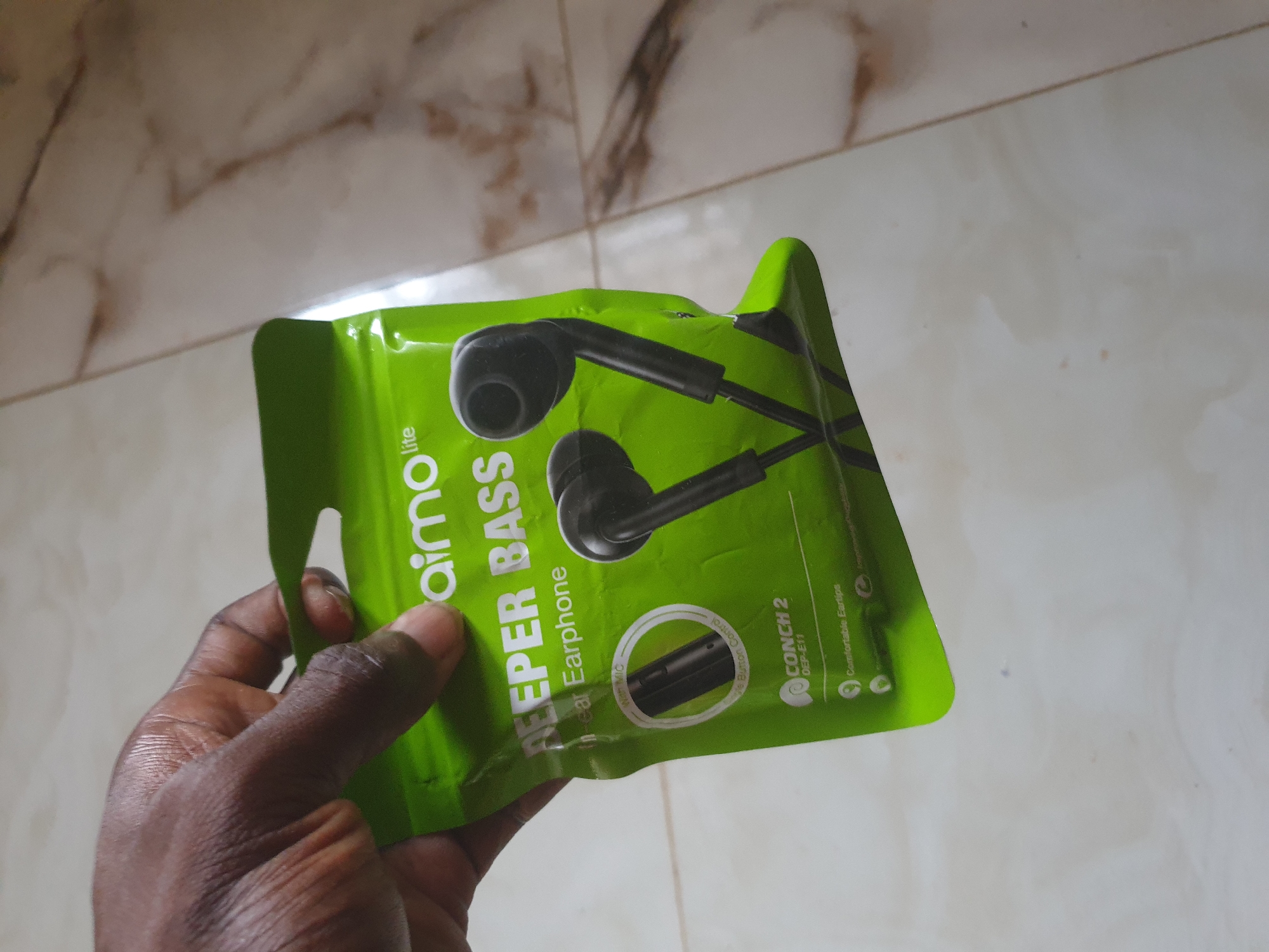 A person's hand, with dark skin, holds a bright green package containing black earphones. The packaging displays the text "aimo lite", "DEEPER BASS", "In-ear Earphone", and "CONCH 2 DEP-E11". A circular graphic on the package shows an earphone with a microphone. The earphones themselves are visible through the plastic packaging. The background consists of a tiled floor with a marbled pattern. The image is taken indoors, with the lighting suggesting an ambient environment.