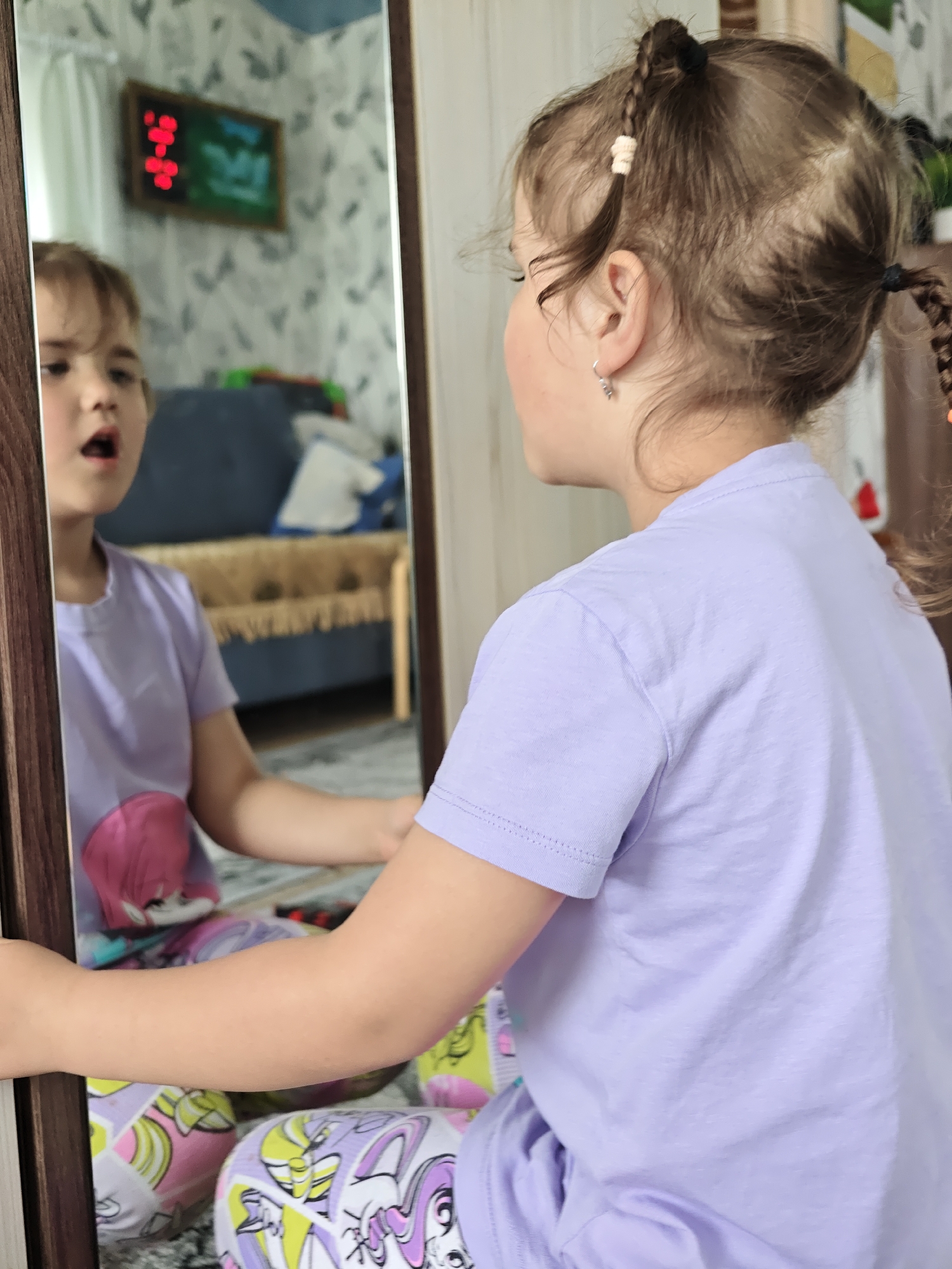 A girl with braided hair and a lavender shirt is looking into a mirror. The reflection in the mirror shows her face, revealing her mouth is open. The background, as seen in the mirror, includes floral wallpaper, a dark blue couch, a wooden table, and a wall-mounted digital clock inside a frame. The scene is located in a room. The location is Уральское, Russia.