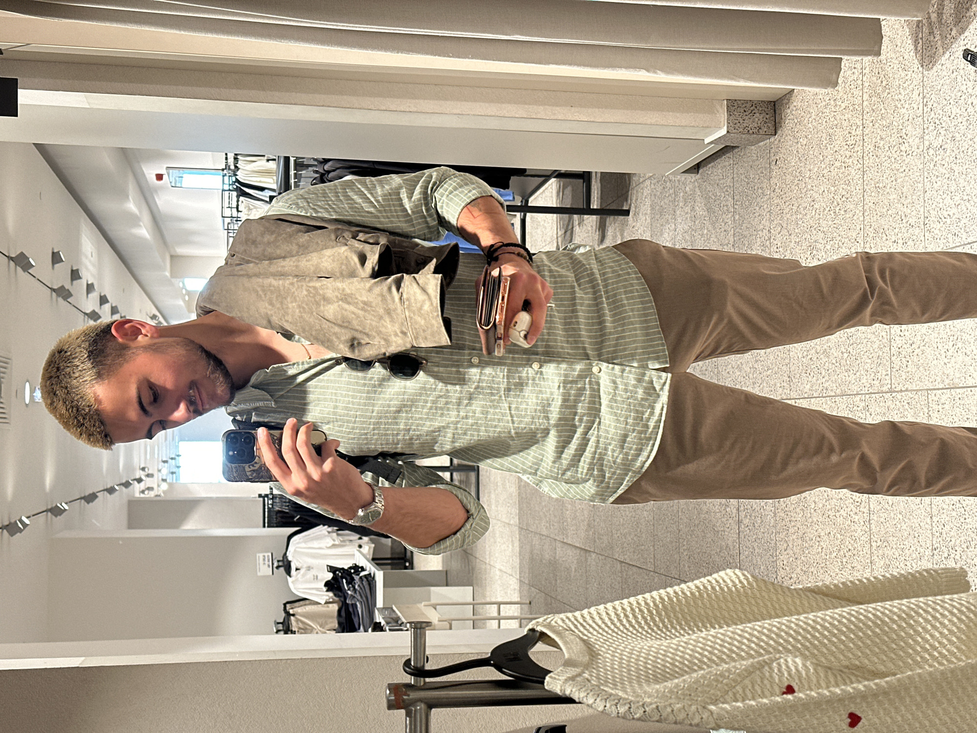 A man takes a selfie in a mirror in a retail store. He is fair-skinned with short, light brown hair and a beard. He wears a light green and white striped shirt with the sleeves rolled up, khaki pants, a brown leather jacket slung over his shoulder, a silver watch, and multiple black bracelets on his right wrist. He holds a cell phone in his left hand, and in his right hand he holds a brown wallet and a white round object. Sunglasses rest on his shirt. The store features racks of clothing, white walls, and a white ceiling with recessed lighting. A white waffle-knit sweater hangs on a rack in the foreground. The floor is covered in a light-colored tile with a speckled pattern. The location is consistent with a retail store in Sofia, Bulgaria.