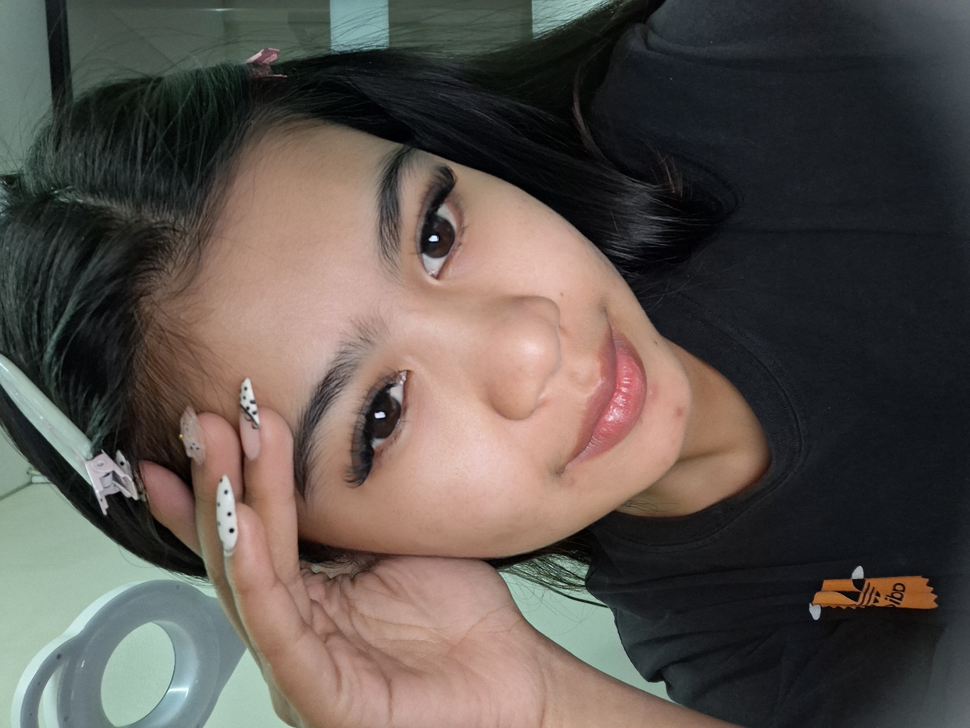 In Kuta, Indonesia, a young woman with dark hair, styled with two pink clips and a white clip, is seen in a close-up portrait. She is wearing a black t-shirt with an orange Adidas logo. Her right hand is raised to her forehead, revealing long, artificial nails with a white and black polka dot design. The background is a plain, light-colored wall with a ring-shaped object visible on the left. The woman has noticeable makeup, including dramatic false eyelashes.