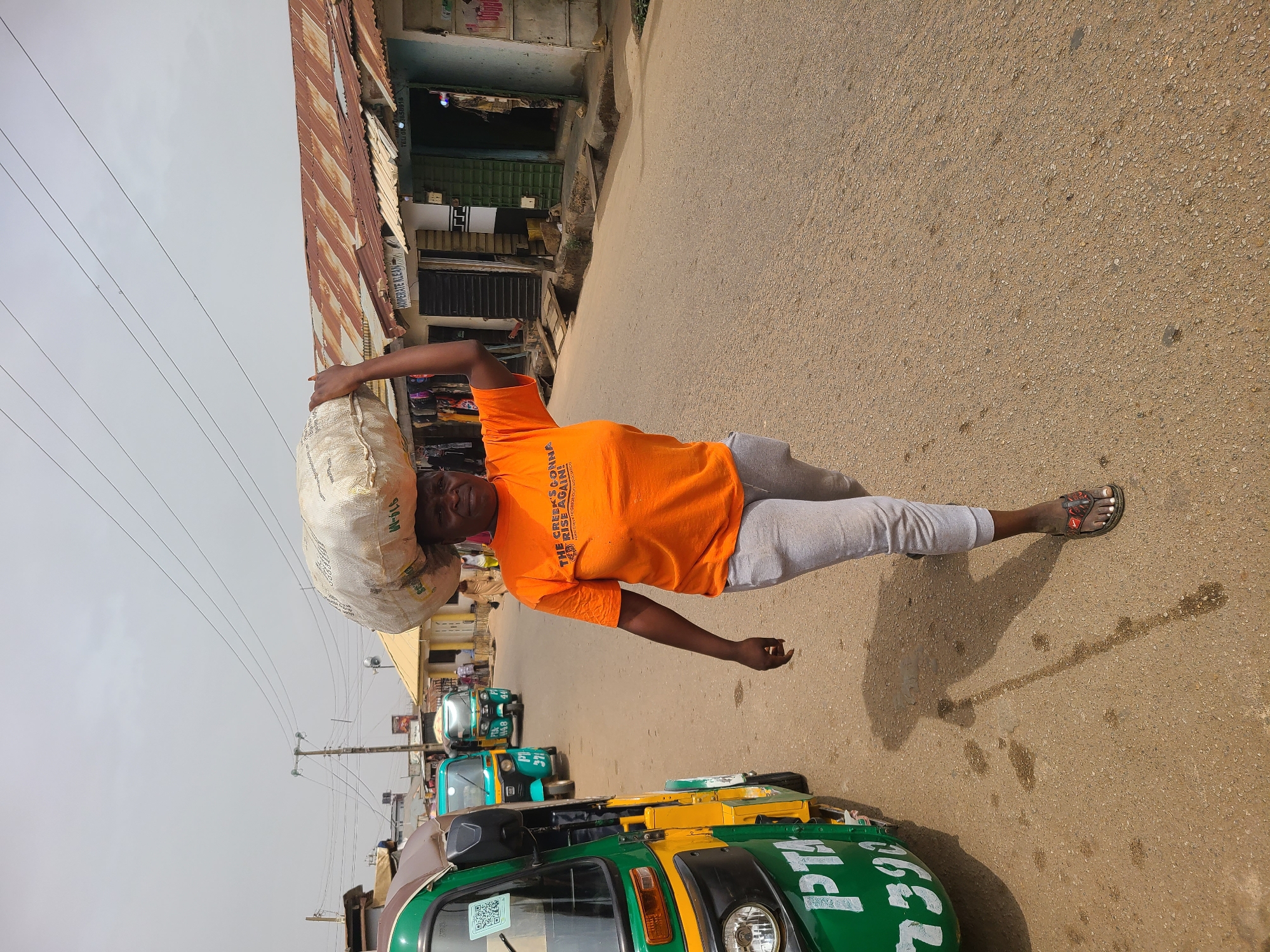 A man in Jos, Nigeria, carries a large, white sack on his head while walking on a paved road. He is wearing an orange t-shirt with text, grey pants, and sandals. In the background are shops and green and yellow auto rickshaws (tuk-tuks). One of the rickshaws has "PT 393 M" written on it. Overhead power lines are visible in the sky.