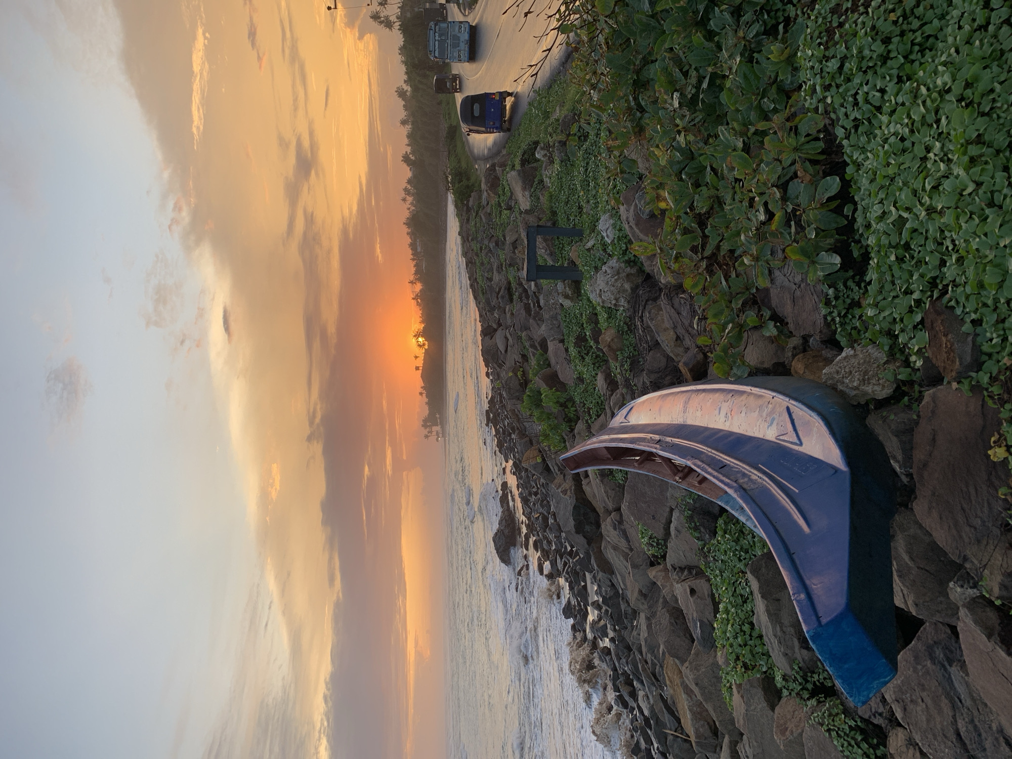 Captured in Kathaluwa, Sri Lanka, the image presents a seaside scene during sunset. The sky transitions from light to dark with prominent orange and yellow hues. The ocean's waves touch the rocky shore, where vegetation is growing. Discarded on the rocks is a blue, possibly vehicle-related, structure. A road with moving vehicles runs parallel to the shore. The scene has no visible people.